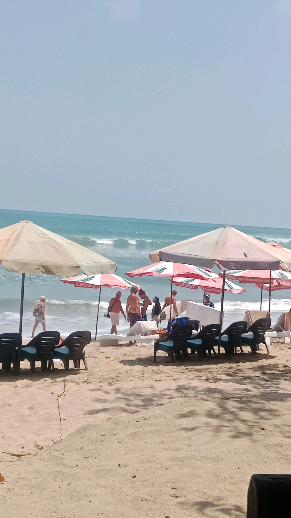 The image depicts a beach scene in Kuta, Indonesia. In the foreground, several lounge chairs are arranged on the sandy beach, some with blue cushions, facing the ocean. Two large beach umbrellas, one beige and another with red and white stripes, provide shade. A group of people are seen near the water's edge, some walking and others standing. One person is carrying a surfboard. The ocean has moderate waves, and a few small whitecaps are visible. The sky is a pale blue with no clouds. The overall impression is a sunny day at a popular tourist beach.