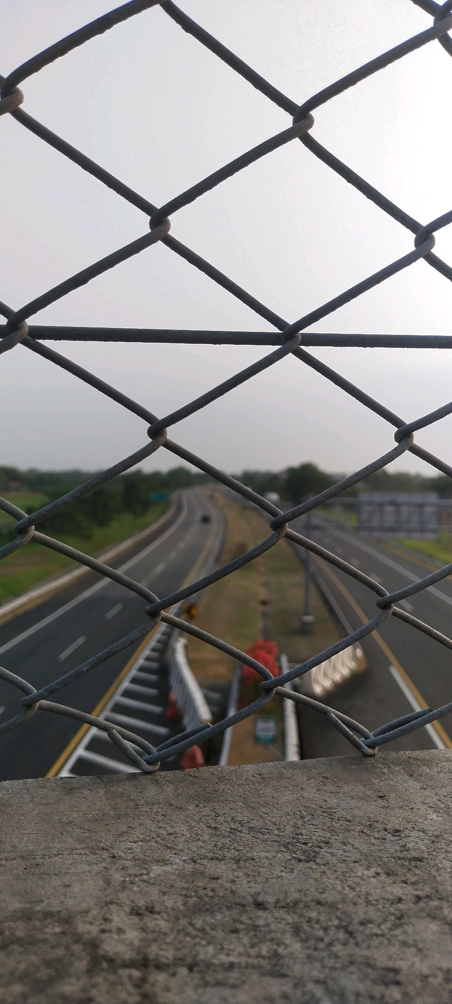 The image is a vertical shot, taken from an elevated position, looking through a chain-link fence at a highway in Desa Sobokerto, Indonesia. The fence's diamond pattern dominates the foreground, with the mesh wires creating a textured barrier. Below the fence, a concrete ledge is visible, adding to the sense of height. The highway stretches into the distance, with multiple lanes marked by white and yellow lines. Cars are sparsely visible on the road, indicating moderate traffic. The surrounding landscape consists of green foliage and trees. The sky is overcast and light, suggesting it is either early morning or late afternoon. The overall impression is one of looking out over a busy transportation artery from a protected vantage point.