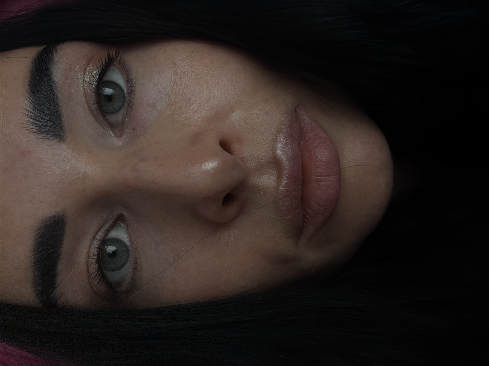 A close-up, portrait-oriented photograph shows a person's face from a low angle, tilted to the right. The person has fair skin, dark hair, and light-colored eyes. The eyebrows are dark and thick. The eyes are wide open, and a slight sheen is visible on the eyelids. The nose is centrally located, and the lips are full and slightly parted. The hair is dark and frames the face, with some strands visible in the background. The overall lighting appears soft and diffused, emphasizing the person's features. There are no indicators that this image was taken in Saint Petersburg, Russia.