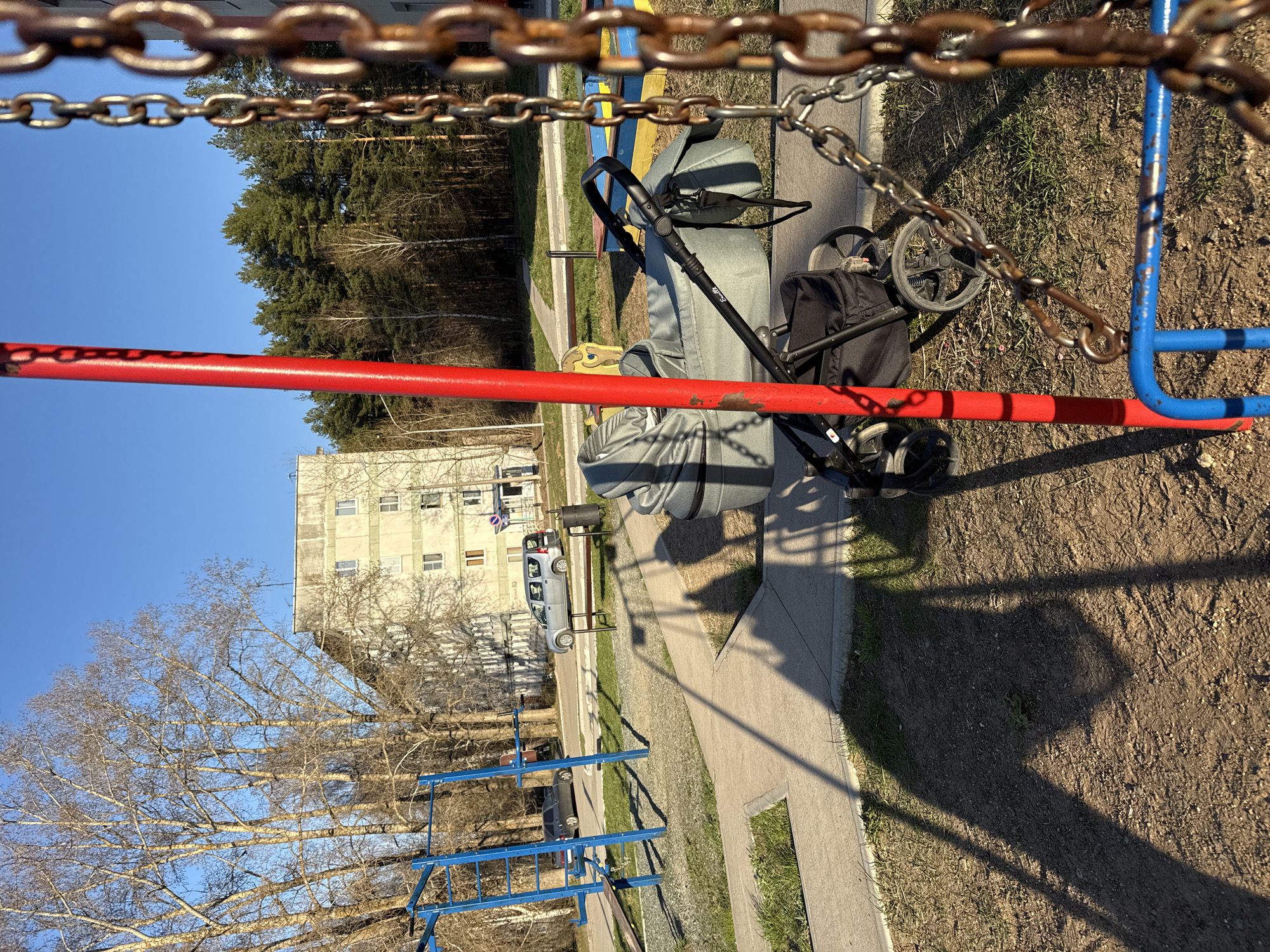 The image depicts an outdoor scene, possibly a playground, in an urban environment. A baby stroller with black wheels and a gray body sits adjacent to a concrete walkway. In the background, there is a large, weathered building with multiple windows. A vehicle is parked on the street in front of the building. In the foreground, sections of playground equipment, including a red bar and blue structures, are visible. Rusty chains hang at the top of the frame, adding an element of enclosure to the view. Bare trees with intricate branching patterns suggest it might be early spring or late autumn. The ground consists of dirt and sparse grass. The sky is clear blue.