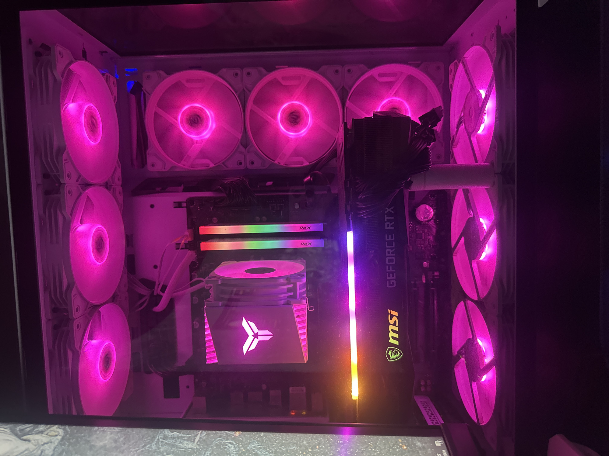 The image shows the interior of a computer case illuminated by pink RGB lighting. Multiple fans are visible, some on the top of the case, some on the side, and some at the bottom. A graphics card, identified by the "msi GEFORCE RTX" logo, is mounted horizontally in the case. Above the graphics card is a CPU cooler with a distinctive logo. RAM sticks with RGB lighting are also visible. The overall impression is of a high-end gaming or enthusiast PC build. The image is not in a city and there is no indication of it being in any specific location.