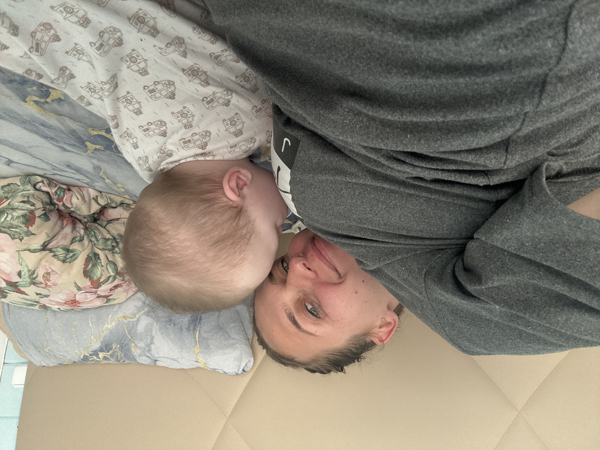 A photograph depicts a woman lying on her back next to a baby. The woman has fair skin, short dark hair, and light-colored eyes. She is wearing a gray sweatshirt. The baby is lying on its side with its head close to the woman's face. The baby has fair skin and light hair. The baby is wearing a white sleeper with a pattern of robots. They are on a bed covered with a light beige quilt. There are several pillows behind the baby, including one with a floral pattern and another with a marble pattern. The photo was probably taken indoors, on a bed in a room in Razdolye, Russia.