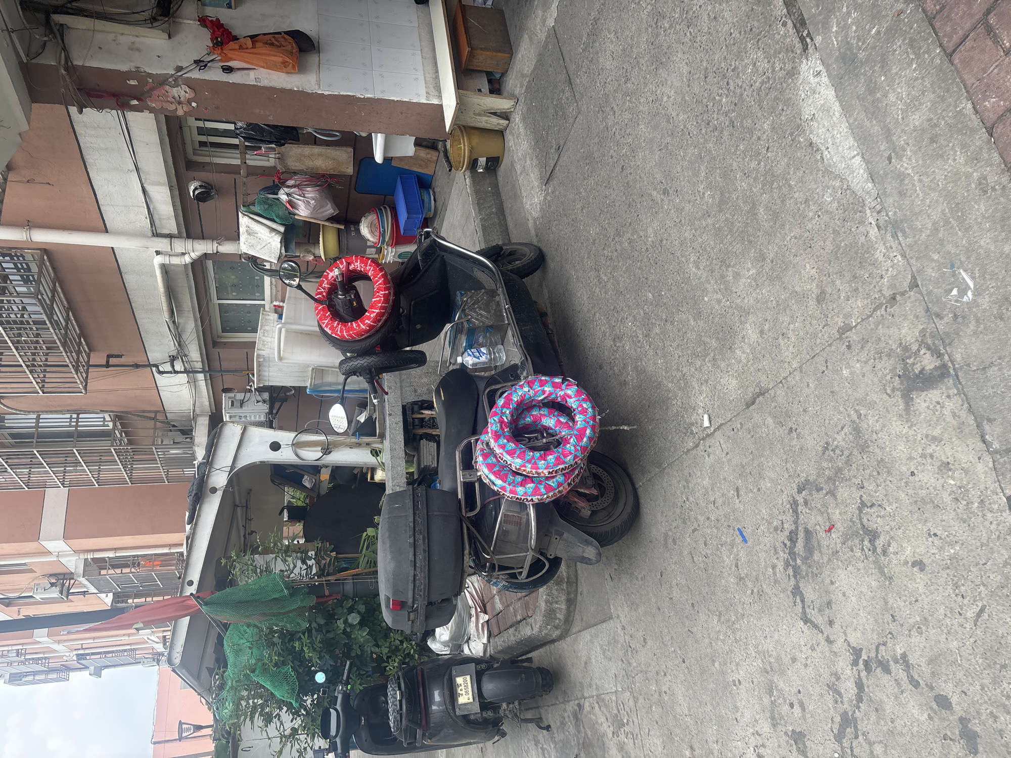 Two dark-colored electric scooters are parked on a concrete sidewalk in front of a building. The scooter on the right is larger and has a black storage box on the back, above which are two circular, inflatable rings, one red and one patterned with pink and blue. The scooter on the left is smaller and has a license plate that reads "0658200". The building behind the scooters has a light brown facade with several windows and air conditioning units. There are green nets hanging from an overhang to the left of the scooters, and various objects, including buckets and bags, are stored on shelves along the side of the building. The ground is cracked and stained concrete. This scene is located on a street within Suzhou Industrial Park, China.