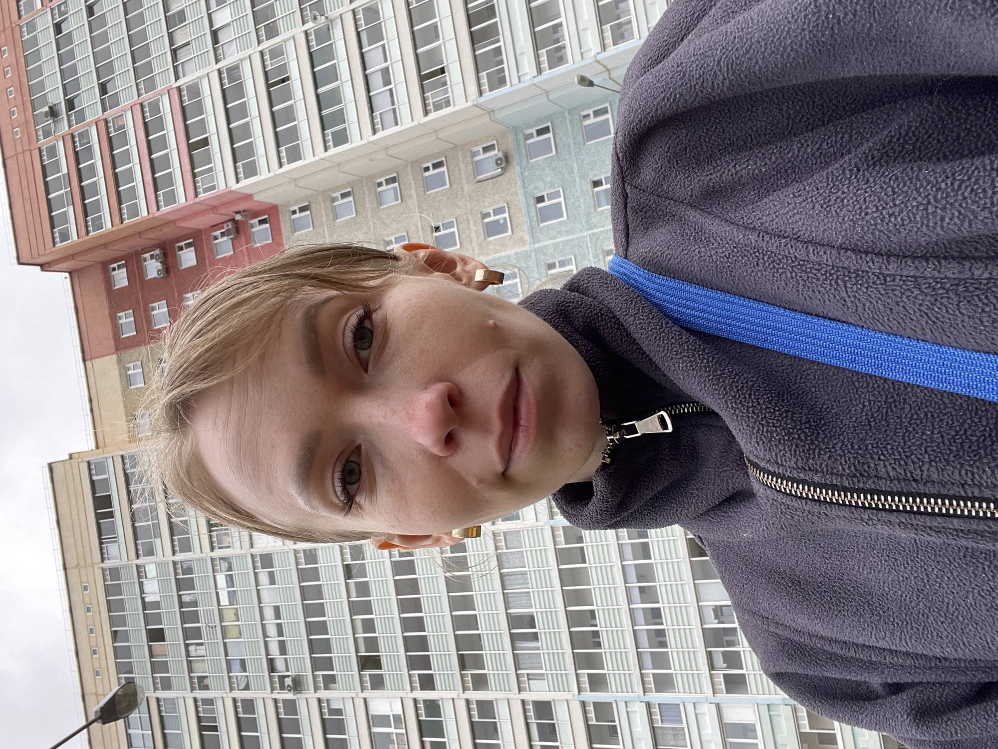 This vertical-format selfie, rotated 90 degrees clockwise, captures a young person in the foreground against the backdrop of a large urban building.
The subject is a young individual with light-colored hair, looking directly into the camera with a neutral expression. They are wearing a dark grey or navy blue fleece jacket with a silver zipper visible on the right side of the frame (when upright). A bright blue strap, likely from a bag, crosses their chest. A simple, gold-colored rectangular earring can be seen on their right earlobe.
The background features a tall, multi-story building, characteristic of an urban environment. Its facade is composed of numerous rectangular windows with light frames, arranged in a grid pattern. Distinct sections of the building are painted in different colors, including a prominent reddish-brown/light pink area, a lighter blue-grey section, and a beige/tan section. Several air conditioning units are mounted beneath some windows. On the far left, part of a street light pole is visible, indicating an outdoor, public setting.
The scene appears to be set during daytime under an overcast sky, providing soft, diffused lighting. The architectural style of the building, with its uniform window grid and distinct color blocks, is suggestive of post-Soviet era construction, consistent with the provided location context of Perm, Russia. No visible text is present in the image.