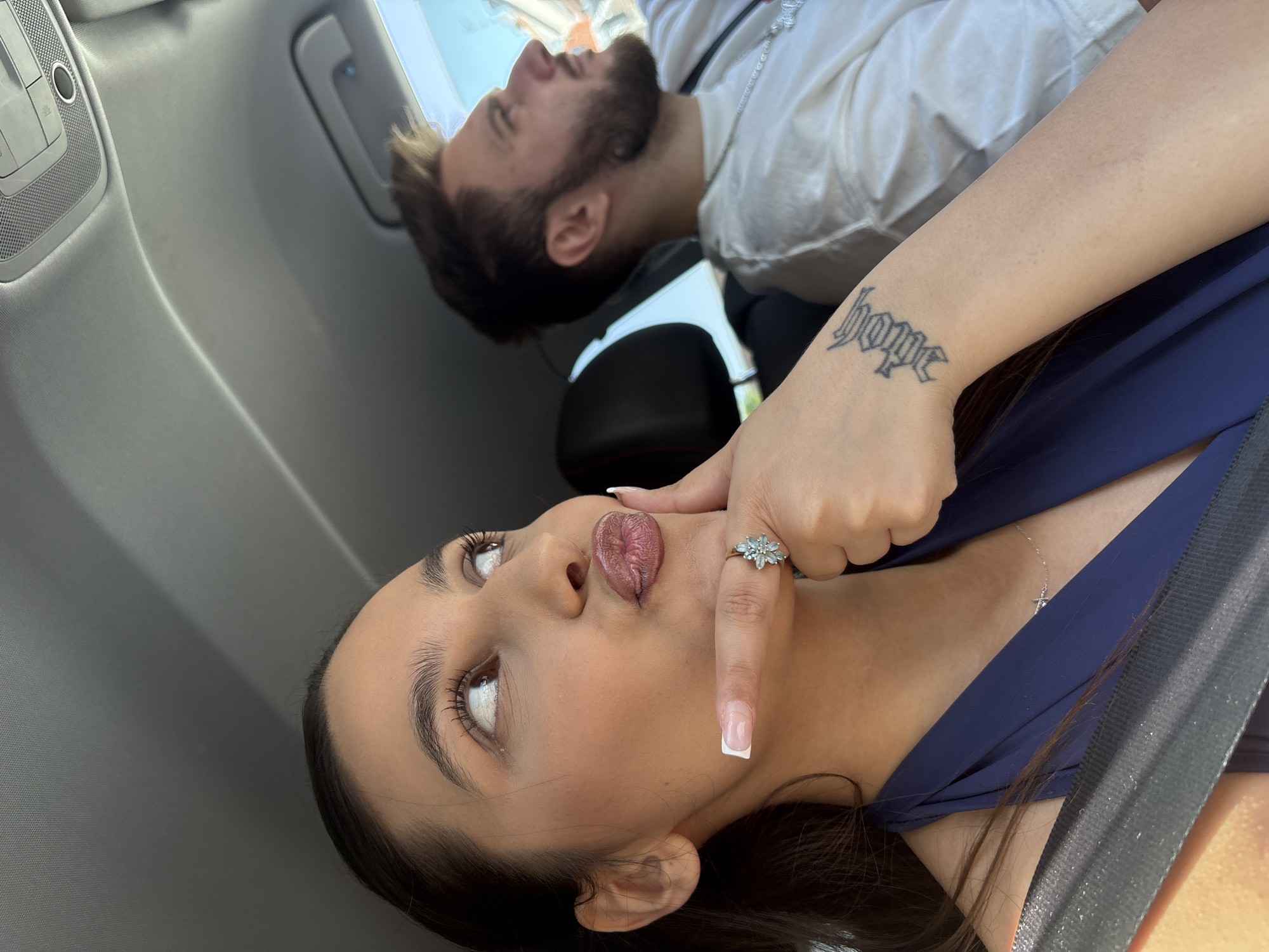 The image shows two individuals in what appears to be the interior of a car. The foreground focuses on a woman with brown hair, fair skin, and brown eyes, positioned at an angle. She is wearing a dark blue top and a necklace with a cross pendant. Her tongue is protruding, and she is holding her index finger against it. A ring is visible on her finger. A tattoo that reads "hope" is visible on the woman's wrist. In the background, a man with a beard and fair skin is lying down, wearing a white shirt and a silver chain. The interior of the car is visible, with gray panels and a black headrest. The location in the city of Mönchengladbach, Germany cannot be determined from the given information.