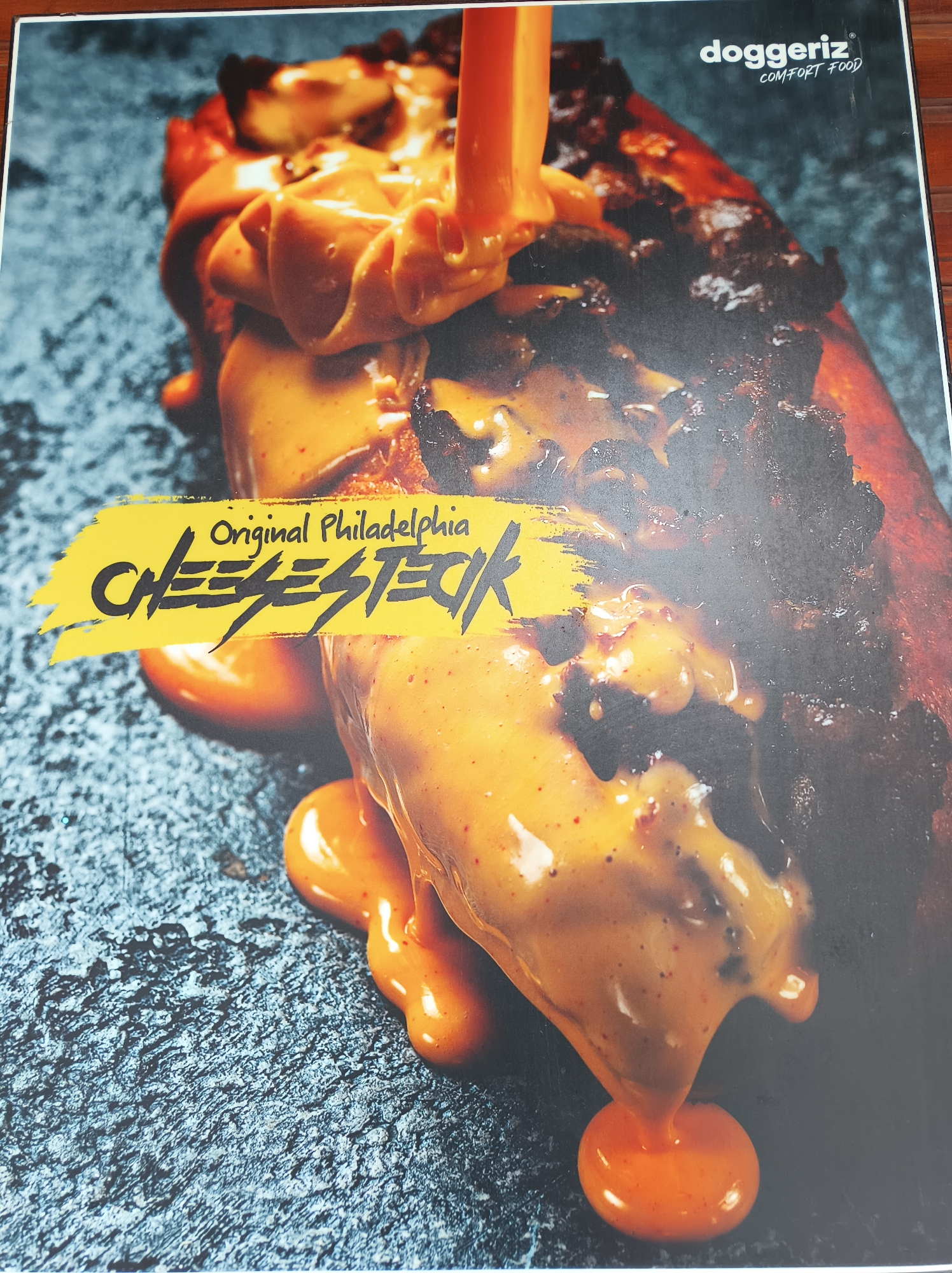 The image shows a close-up of an "Original Philadelphia Cheesesteak" from "doggeriz COMFORT FOOD". The cheesesteak is heavily covered in melted cheese sauce, which is also being poured onto it. The background is a dark, textured surface.