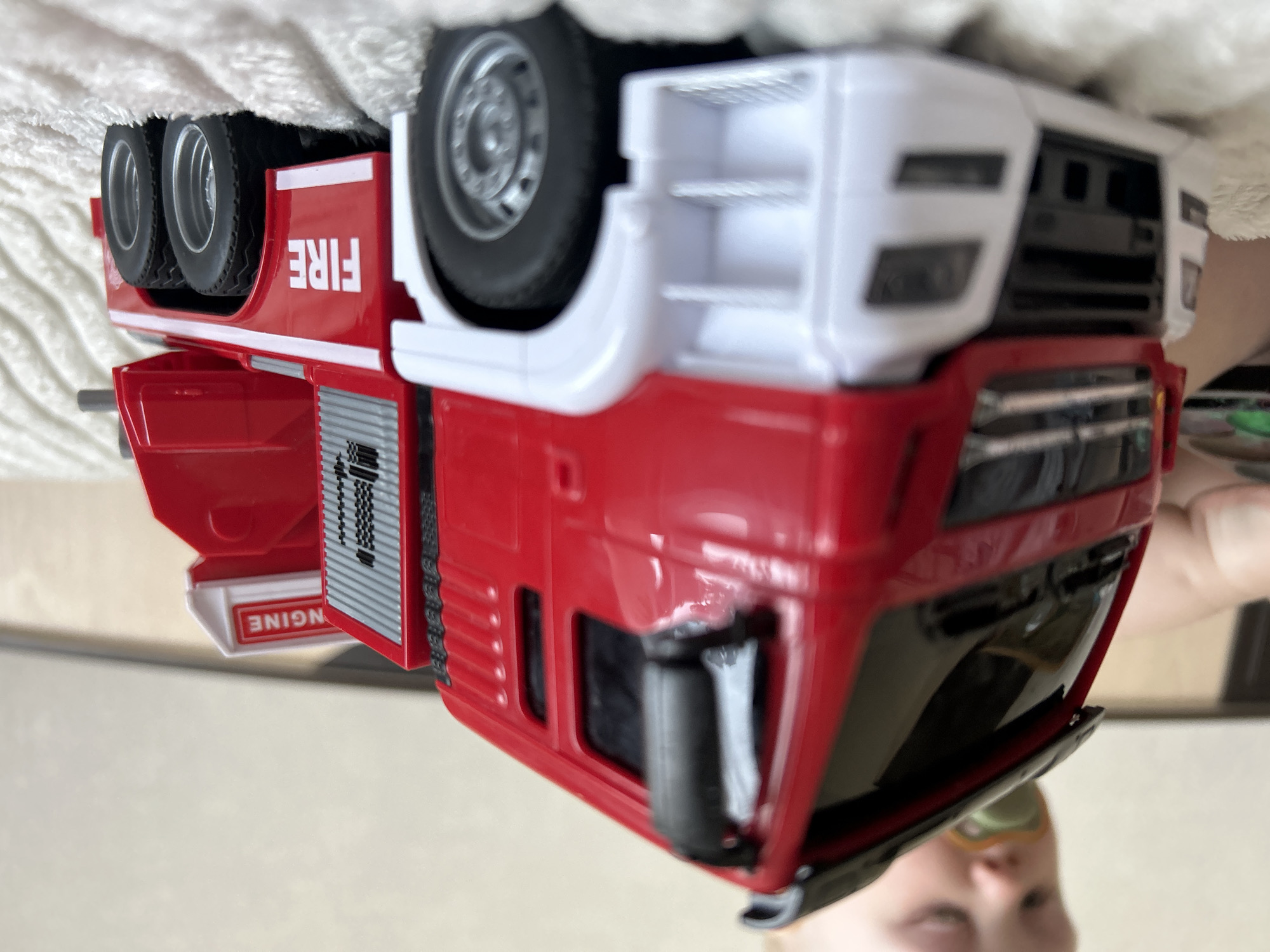 A close-up, upside-down view features a toy fire engine. The toy is red and white, with the word "FIRE" printed on its side. Its wheels are visible, and part of a label reading "ENGINE" is also seen. A child's face is partially visible in the bottom of the frame. The background appears to be a neutral-toned surface. There is no indication of Chaikovsky, Russia in the image.