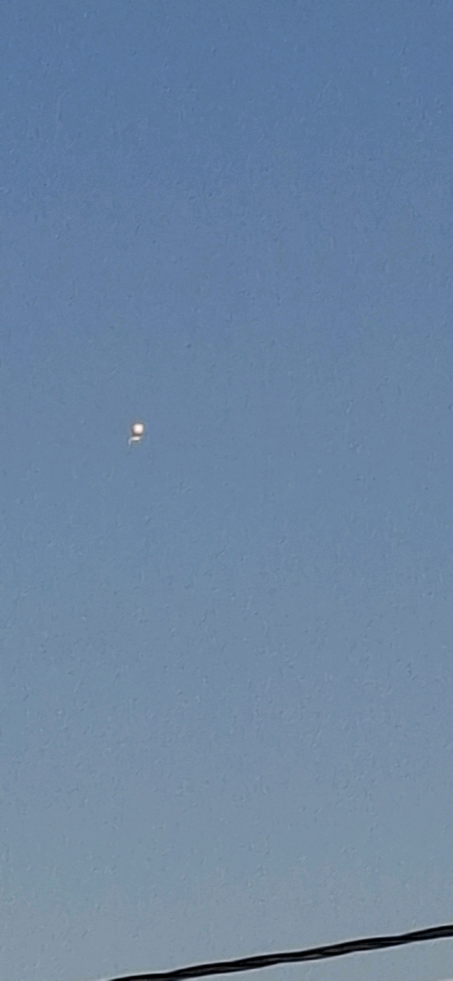 The image captures a bright, rectangular object in a clear blue sky. The object is small and appears to be luminous, with a distinct white glow. Below the sky, a dark, horizontal line, likely a power line, is visible at the bottom of the frame. The overall scene suggests an outdoor environment, potentially in Yerocham, Israel, given the context.