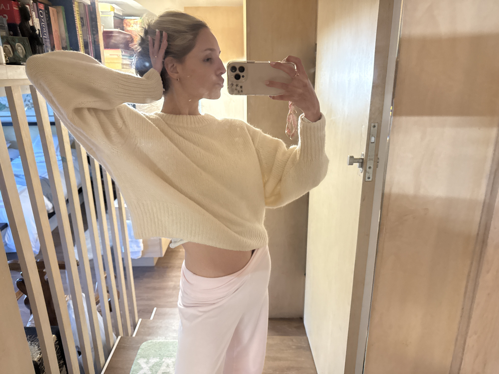 A young woman with blonde hair pulled up is taking a mirror selfie in what appears to be a modern apartment. She is wearing a cream-colored knit sweater and light pink lounge pants. Her left arm is raised, with her hand touching the back of her head, and her right hand is holding a smartphone to take the picture. She is looking at her reflection and puckering her lips. The background shows a bookshelf filled with books, a wooden staircase, and a door with a visible latch. The overall environment is clean and well-lit, suggesting a residential setting. There is no discernible evidence to place this image in Ciudad de México, Mexico.