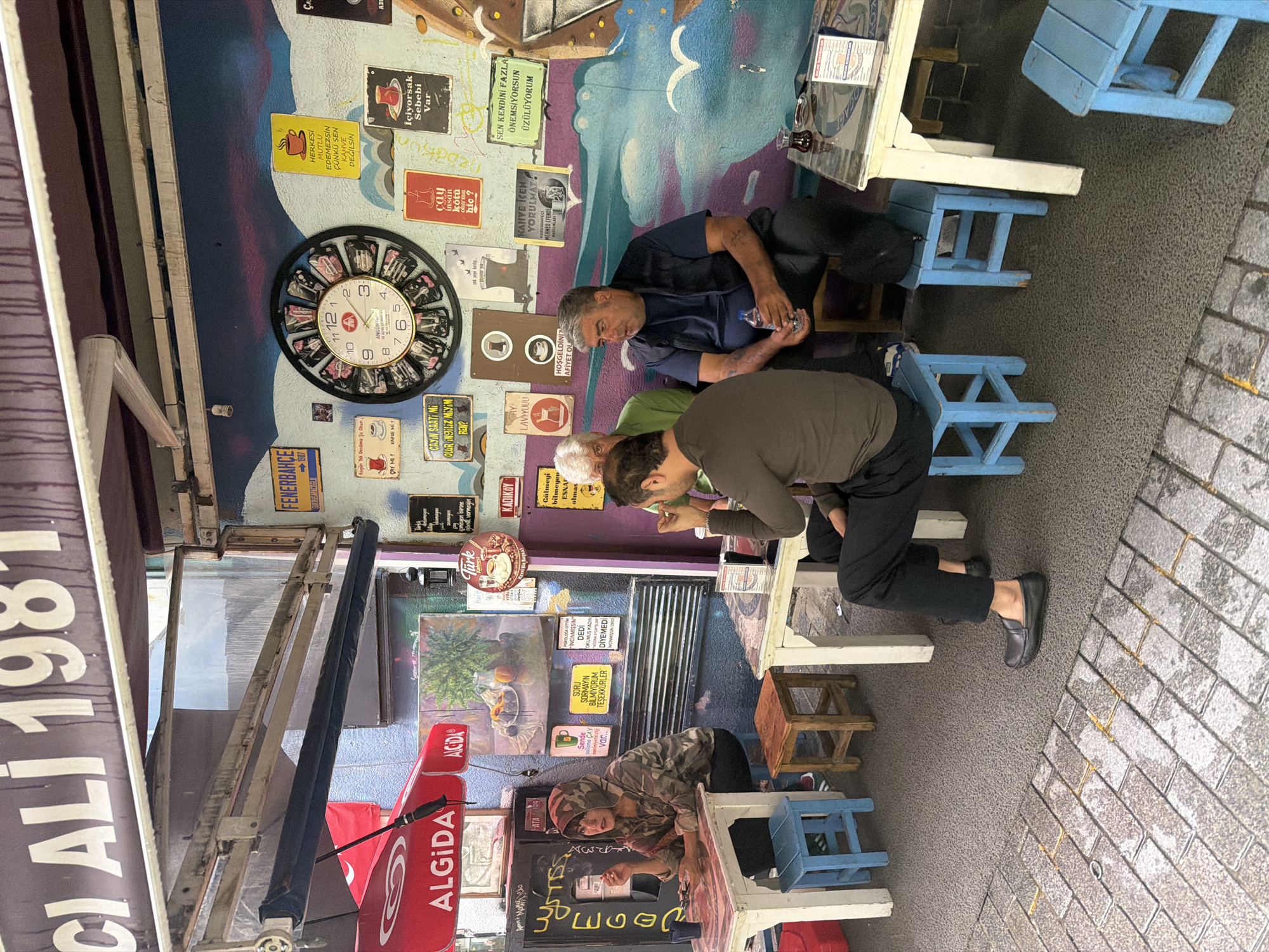 The image depicts an outdoor seating area of a cafe or tea house in Kadıköy, Turkey. The walls are decorated with a vibrant mural featuring clouds, birds, and various signs, some in Turkish. A large, retro-style clock hangs prominently on the wall. Several individuals are seated at small tables, some under an awning. One woman, wearing a camouflage hooded jacket, sits alone at a table on the left, holding a cigarette. In the center, two men and another individual are seated. One man in a green shirt appears to be engaged in conversation, while another in a dark vest holds a bottle of water. On the right, a man in a dark shirt is seated. The seating consists of small, colorful wooden stools and tables. An Algida ice cream umbrella is visible on the left. The ground is paved with cobblestones and a textured surface. The overall scene suggests a casual, communal gathering spot.