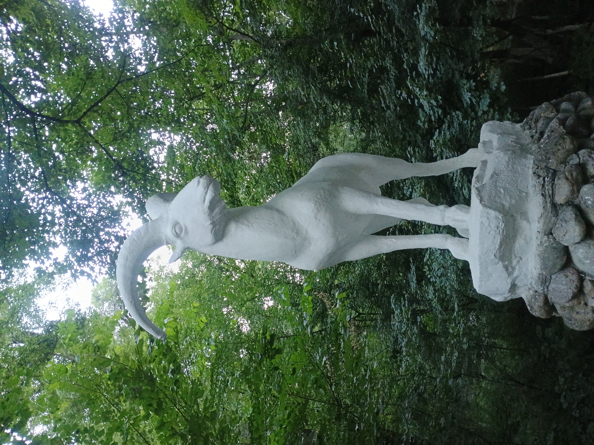 A full shot, low-angle, outdoor photograph captures a white, concrete statue of a ram positioned on a rock formation in the city of Gusar, Azerbaijan. The ram statue stands on its four legs, facing the viewer's left, with its head tilted slightly upwards. Its left horn curves prominently upwards and to the left, while its right horn is partially obscured. The statue is set against a backdrop of lush green trees and foliage, with dappled sunlight filtering through the leaves from above. The rock formation beneath the statue consists of rough, natural stones, suggesting an outdoor, possibly park-like setting. The lighting suggests it is daytime, with overcast conditions contributing to the soft light. There are no people or animals present in the image.