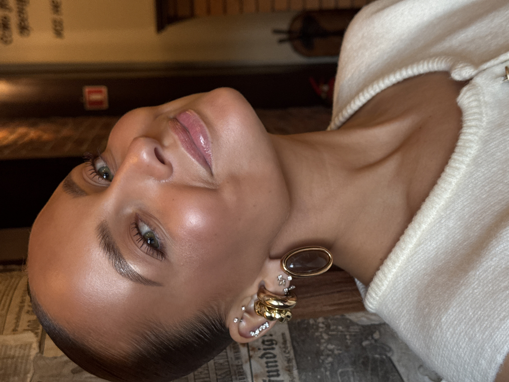 A close-up portrait of a woman with light skin and green eyes, her head tilted slightly upwards and to the right. She has dark, slicked-back hair, styled neatly away from her face. Her makeup is subtle, with a focus on glowing skin, defined eyebrows, and a glossy pink lip. She is wearing multiple gold hoop earrings on her left ear, some adorned with small rhinestones, and a larger oval earring with a dark brown stone. She is also wearing a white knitted sweater with a ribbed neckline. The background is blurred, with hints of a brick wall on the right and a textured, possibly newspaper-like surface on the left. The lighting suggests an indoor setting with warm tones. The specific location in Chaikovsky, Russia is not determinable from the visual information provided.