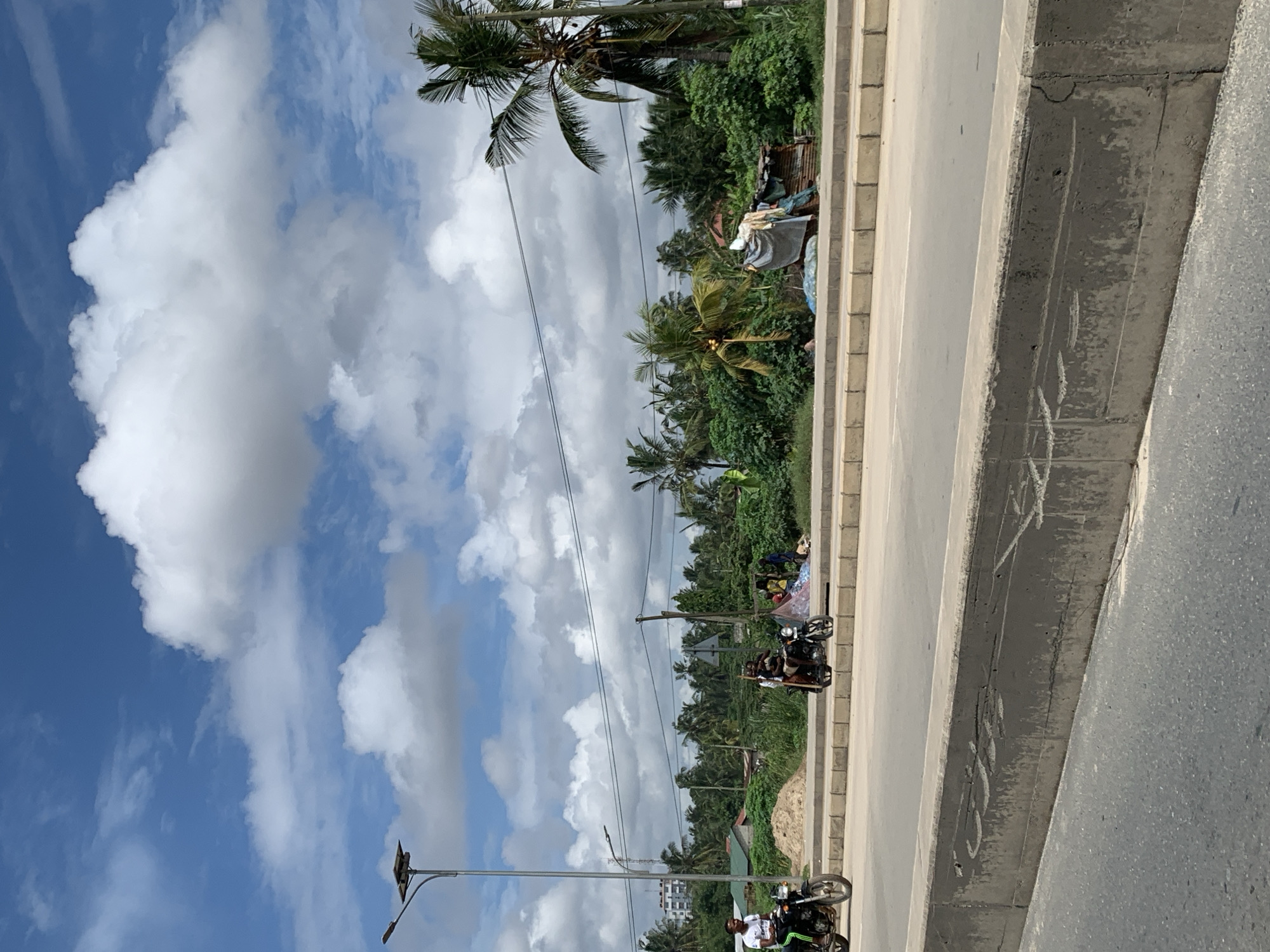 The image depicts an urban scene in Dar es-Salaam, Tanzania. A road runs horizontally across the frame, bordered by a concrete curb. On the left side of the road, vegetation including palm trees is visible. Two motorcycles with riders are on the road. Above the landscape, a bright blue sky is filled with puffy white clouds. Power lines stretch across the sky. A streetlight pole stands in the foreground. Evidence of urban construction can be discerned.