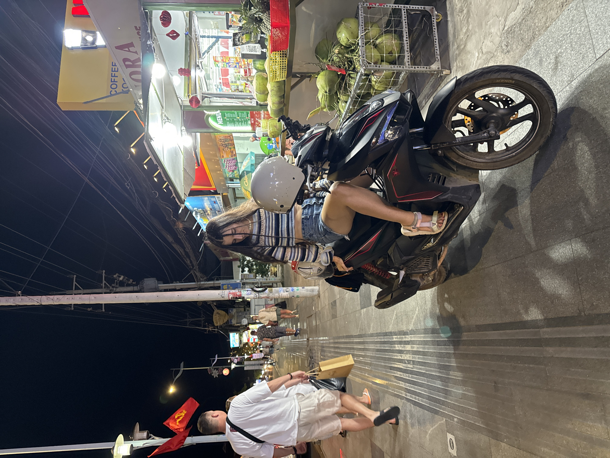 A motorcycle is parked on the sidewalk. A person with long dark hair is sitting on the motorcycle wearing a helmet, a striped top, and denim shorts. There is a small building to the left, with a store with fruit for sale. People are walking along the sidewalk. In the distance, more buildings and lights can be seen. The image appears to be taken at night in Mũi Né, Vietnam. A man in a white shirt walks past the motorcycle.