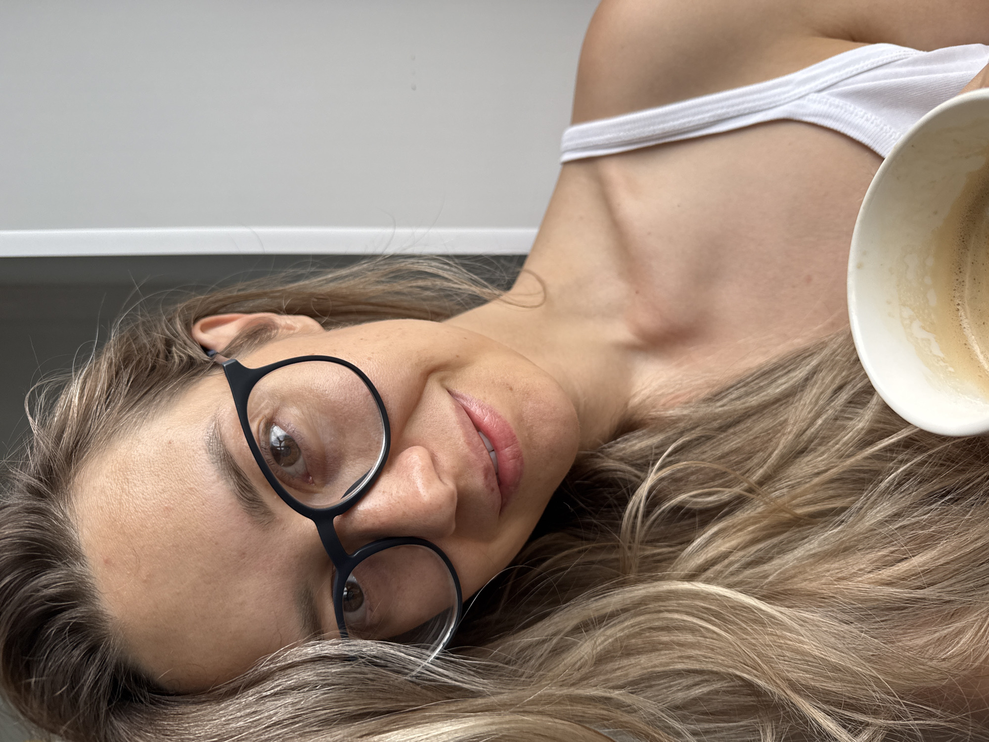 A woman wearing black-rimmed glasses, a white tank top, and a slight smile is holding a cup of coffee. Her blonde hair is partially covering her face and shoulder. The background is a plain gray wall and a white doorframe, suggesting an indoor setting. The image is a close-up of her face, with the coffee cup in the foreground. There is no indication of the city of Chaikovsky, Russia, in this image.