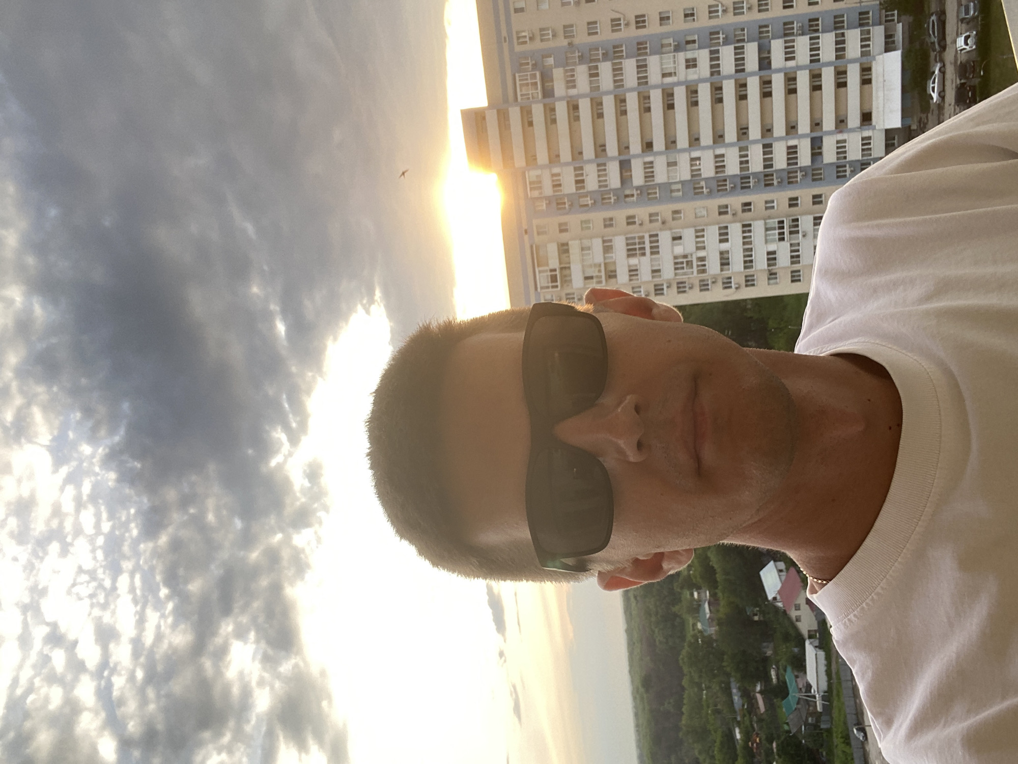A man is taking a selfie outdoors in front of a tall apartment building during sunset. He is wearing a white t-shirt and black sunglasses. The sun is setting behind the building, casting a warm glow on the sky and clouds. The background shows a neighborhood with houses and trees. The image is taken from a high vantage point, likely from a window or balcony of a building in Novokuybyshevsk, Russia.