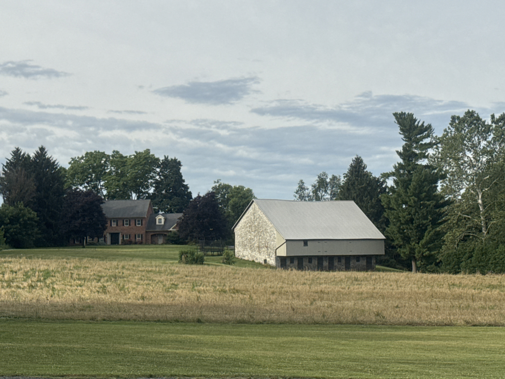 The image shows a rural landscape under a cloudy sky. In the mid-ground, there is a large, light-colored barn with a metal roof and stone facade. To the left of the barn, a two-story brick house with a dark roof is visible, nestled amongst trees. The foreground consists of a manicured green lawn, transitioning to a field of dry, golden grass. The background is dominated by a dense line of various trees, including evergreens and deciduous varieties. The overall scene suggests a quiet, possibly agricultural setting.