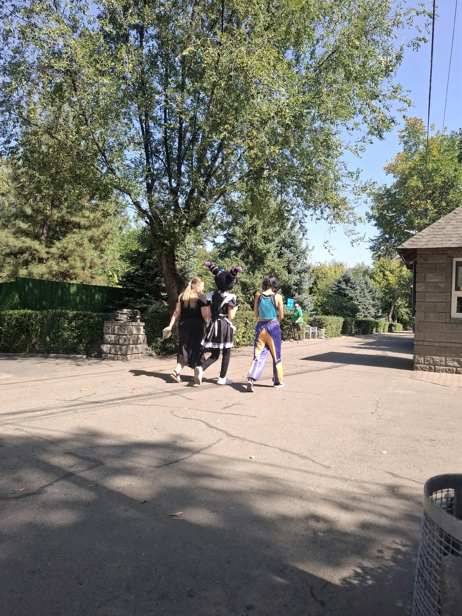 In Almaty, Kazakhstan, three individuals are walking away from the camera on a paved path, which appears to be within a park or outdoor recreational area. The person in the middle is wearing a black and white maid outfit with a purple striped hat. To their left, a woman with blonde hair is wearing a black dress and holding a light-colored object. To the right, a person in a turquoise tank top and purple and yellow striped pants walks alongside. Behind them, a green bush wall lines a path, and further back, various trees and foliage are visible under a clear blue sky. A stone structure, possibly a decorative element or a bench, is positioned to the left of the path. A building constructed of stone blocks is visible to the right, with a pitched roof. The ground is dappled with shadows from the surrounding trees.