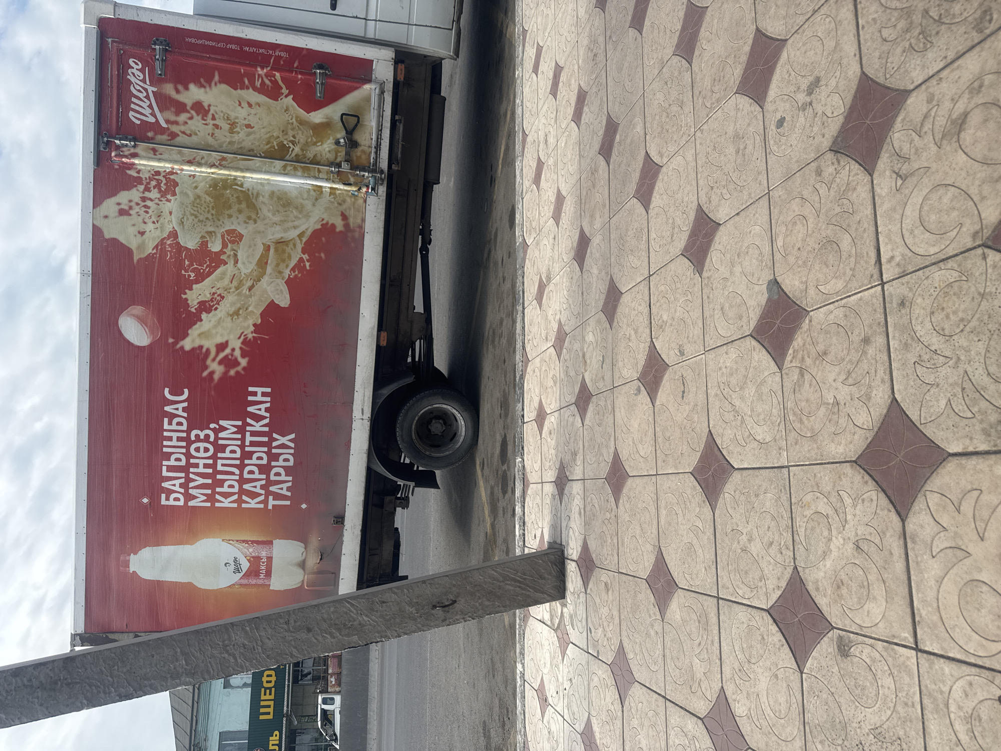 An outdoor, eye-level shot depicts the side of a red truck with a large advertisement on its rear panel. The advertisement features a white bottle with a red label, a splash of yellow liquid, and white text in a non-Latin script. The truck is parked on a street next to a sidewalk with patterned tiles. A concrete pole stands vertically in the foreground, partially obscuring the truck. In the background, buildings and a cloudy sky are visible.