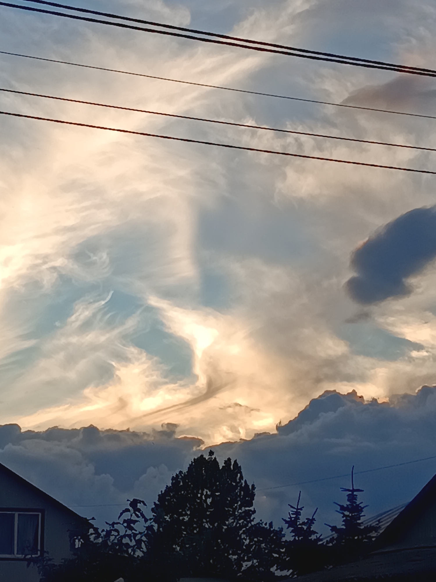 This is a photograph of a sky filled with dramatic clouds, possibly at sunset or sunrise. There are four dark power lines stretching horizontally across the upper portion of the frame. In the lower half of the image, the silhouettes of rooftops, trees, and evergreens are visible against the sky. The sky displays a gradient of colors, with bright, illuminated clouds near the horizon and darker, more ominous clouds above. The presence of power lines and residential structures suggests an urban or suburban setting, likely within the city of Novaya Derevnya, Russia.