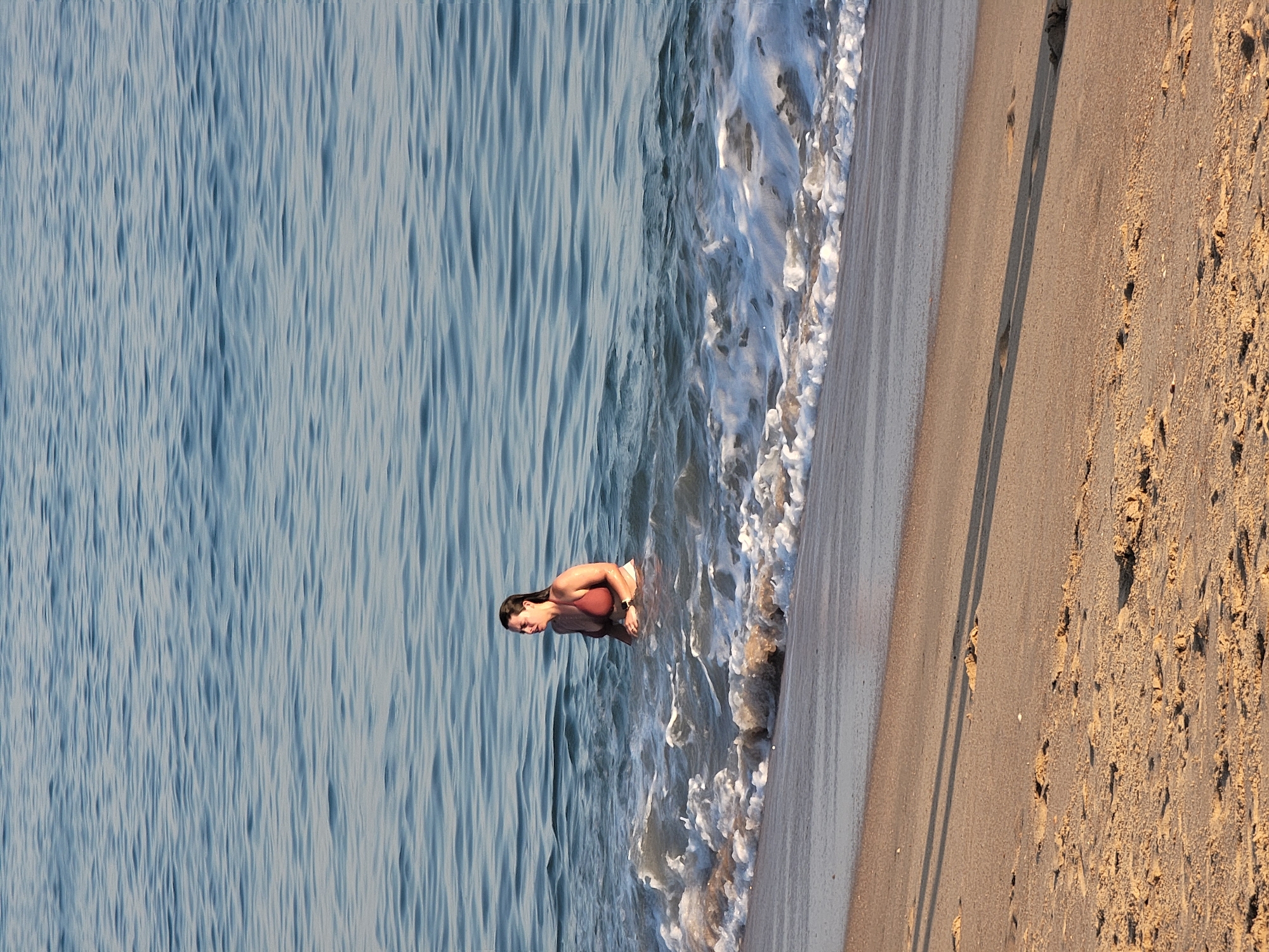 A medium shot captures a woman wading in the ocean at the edge of a sandy beach. The woman, viewed from the side, is submerged in the water up to her chest and is looking to her right. She has dark hair, which appears wet, and is wearing a reddish-brown bikini. The water is a calm blue with gentle waves lapping around her, creating a foamy white edge where the water meets the shore. The beach is composed of light brown sand, with subtle shadows cast across it, suggesting it is either early morning or late afternoon. The scene takes place on a beach in Quarteira, Portugal, with the tranquil ocean and sandy shore as the primary environmental elements.