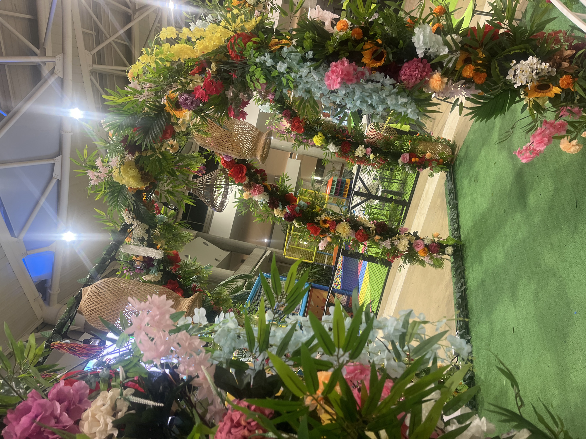 The image shows an elaborate floral archway, possibly a decorative entrance or photo backdrop. The arch is densely covered in a variety of flowers in reds, yellows, pinks, whites, and blues. Woven baskets are suspended from the structure. The arch is situated indoors.
Beyond the archway, there is a glimpse of an indoor playground with colorful elements like nets and slides. The ground around the arch is covered in green artificial turf.
The location is Pampatar, Venezuela. However, there is no specific information related to the city within the image itself.