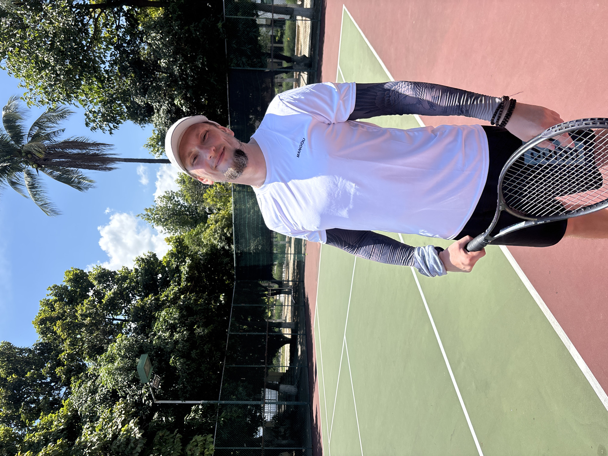 A man in athletic wear stands on a tennis court holding a tennis racket. He is wearing a white t-shirt, black shorts, a white cap, and long-sleeved arm coverings with a tattoo design. The tennis court is green with red out-of-bounds areas and white lines. In the background, there are trees, a fence, and a palm tree, indicating an outdoor setting likely in a tropical or subtropical region. The sky is blue with scattered clouds. Based on the presence of lush tropical vegetation and the style of the tennis court, this image could have been taken at a tennis facility within or near the city of Nha Trang, Vietnam, known for its coastal environment and surrounding greenery.