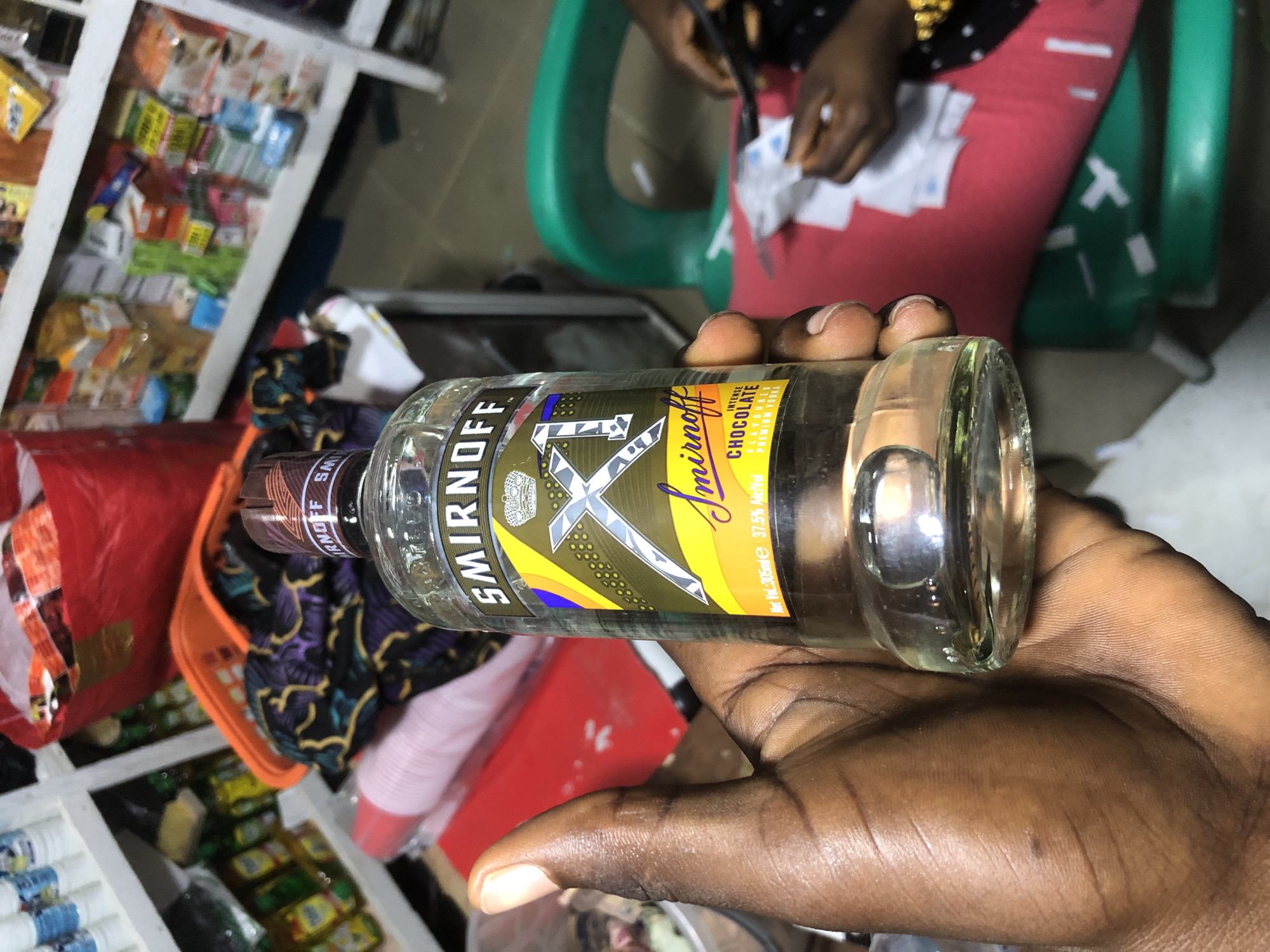 A person's hand is holding a bottle of Smirnoff Ice Triple Distilled Vodka, Chocolate flavor. The bottle is clear glass with a label featuring a gold and silver "X1" design, a crown, and the Smirnoff logo. The label also indicates "Net Vol: 305ml" and "37.5% Alc/Vol". The background is blurred but suggests an indoor environment, possibly a shop or market stall in Ado, Nigeria. Shelves stocked with various products are visible, along with red and orange containers, and a green plastic chair. Another person's hands can be seen in the background, seemingly engaged in an activity with some white paper items.