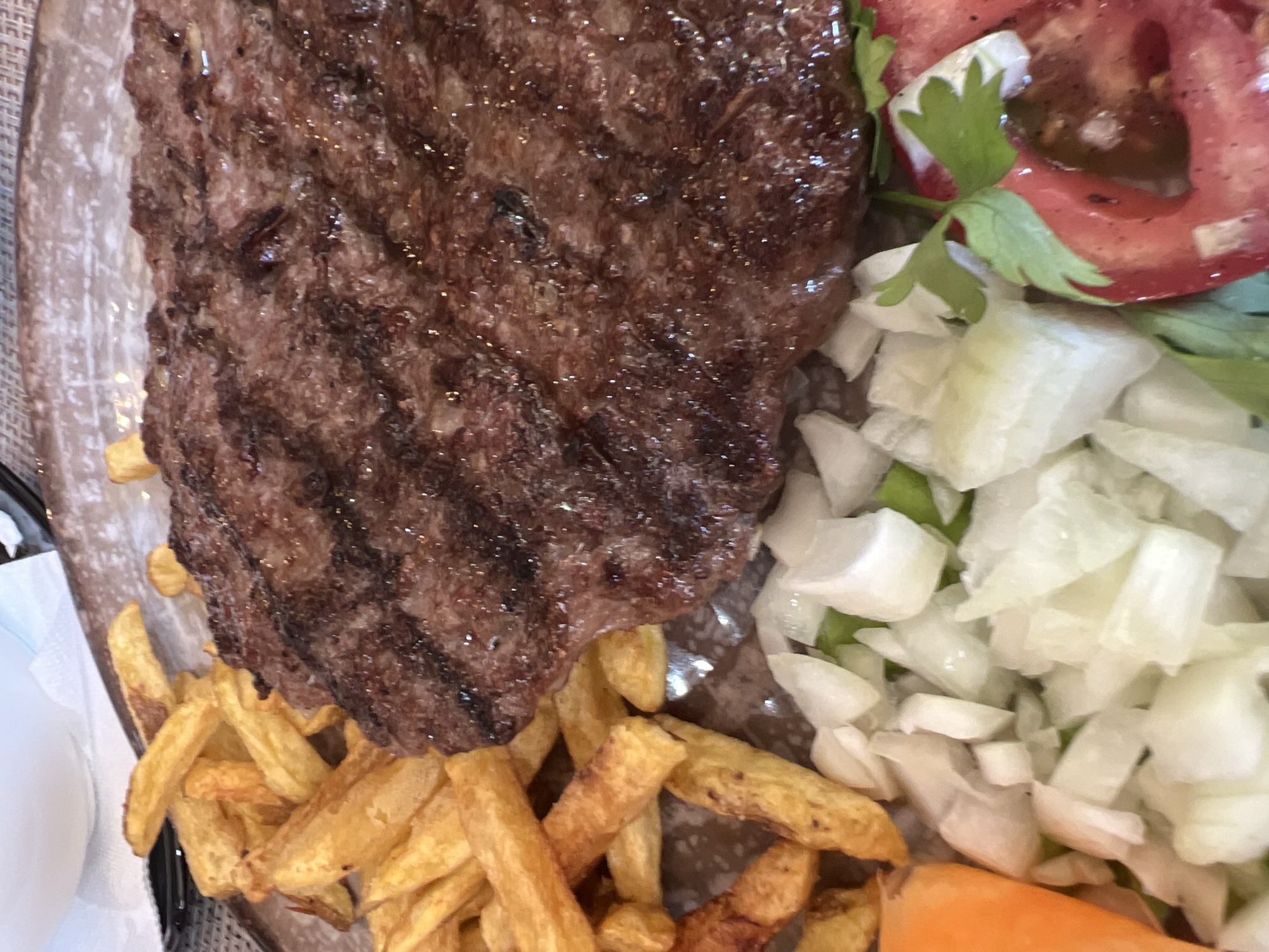 The image shows a close-up view of a meal served on a patterned plate. The meal consists of a large, grilled patty, golden-brown French fries, and a salad of chopped white onions and tomato wedges garnished with parsley. A portion of an orange carrot is visible on the left side of the plate. The lighting suggests an indoor setting, possibly a restaurant. This dish is a typical offering at many restaurants in Budva, Montenegro, a popular coastal city known for its seafood and grilled meats.