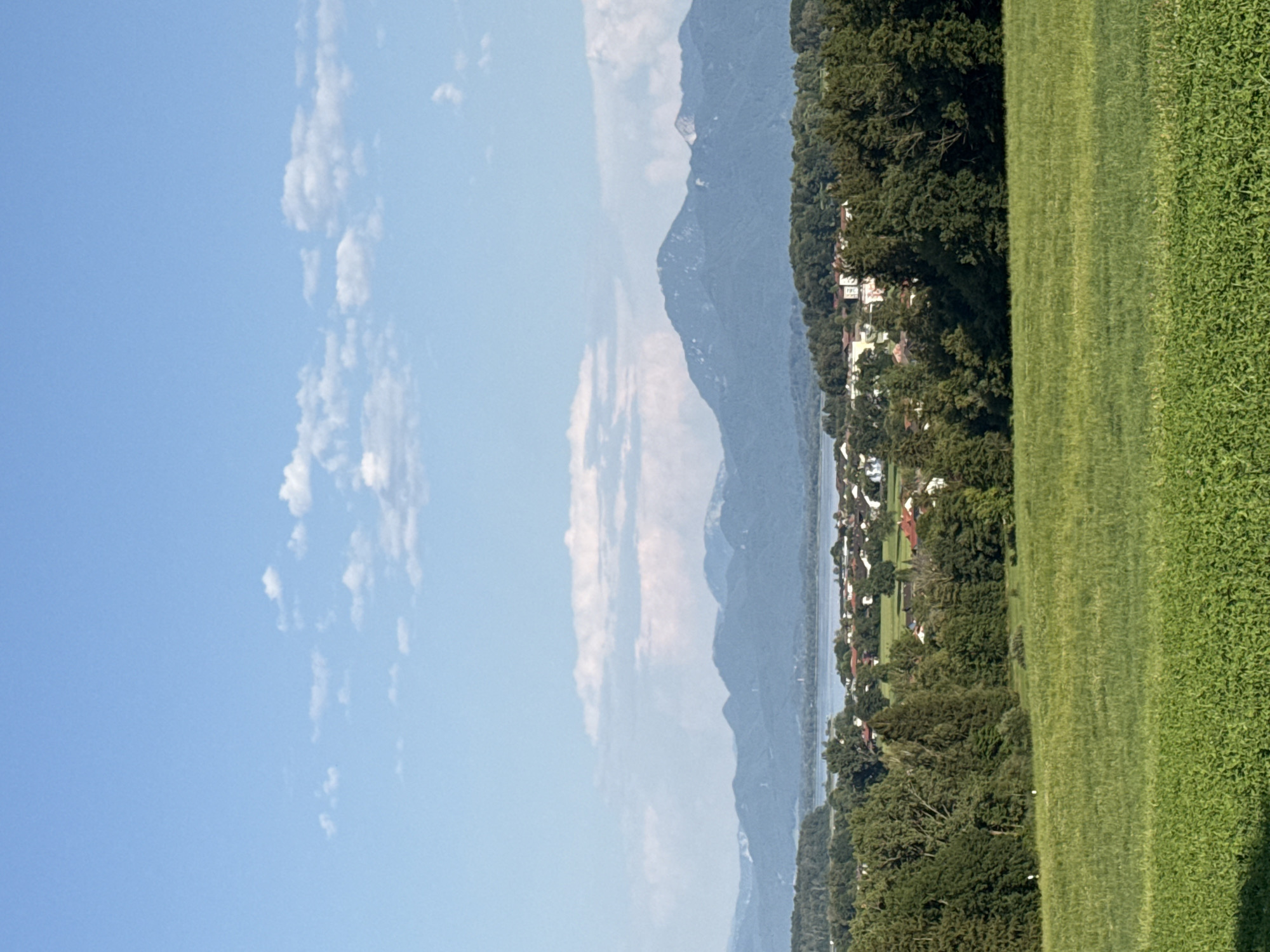 The image captures a picturesque landscape from a slightly elevated vantage point, likely on the outskirts of Rimsting, Germany. In the foreground, a vibrant green, mown meadow dominates, its texture suggesting a well-maintained agricultural field. Beyond the meadow, a dense line of lush green trees forms a natural boundary, with varied shades and textures indicating different tree species.
Nestled behind the trees is a charming village, characterized by a cluster of red-tiled roofs and light-colored buildings, indicative of Bavarian architecture. The village appears to be situated on the gentle slope leading down to a tranquil lake, its surface reflecting the clear blue sky. In the far distance, a majestic mountain range stretches across the horizon, its peaks softly outlined against the pale blue sky. Scattered white clouds drift lazily across the sky, adding a touch of dynamism to the serene scene. The overall impression is one of peaceful rural life, bathed in the warm glow of a summer day.