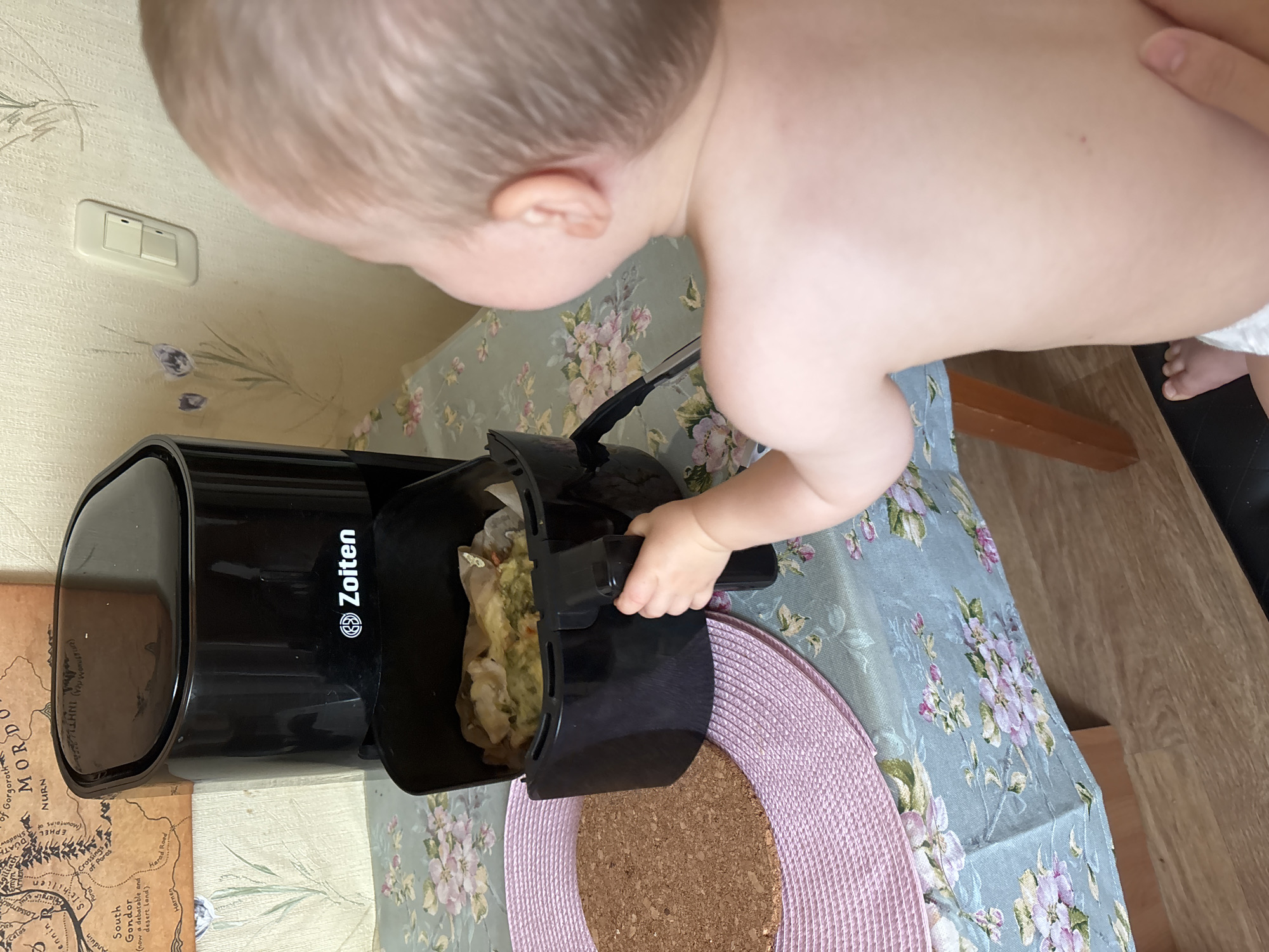 An infant, seen from behind, reaches a hand toward the basket of a black air fryer positioned on a table covered with a floral-patterned tablecloth. The air fryer basket contains food. A cork trivet with a pink placemat is to the left of the air fryer. In the background, a map of Middle-earth is partially visible, attached to a wall with floral wallpaper. A light switch is also present on the wall. The scene appears to be indoors, likely in a residential setting within Naberezhnye Chelny, Russia.