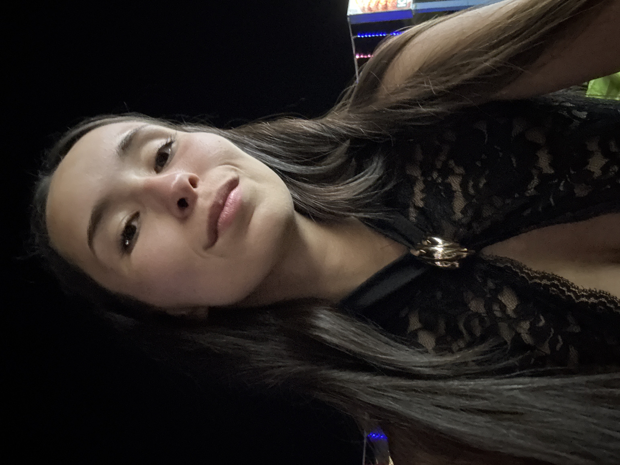 A close-up, low-angle, outdoor selfie at night features a young woman with long, dark hair. She is wearing a black lace top with a gold clasp at the neckline. Her face is illuminated, with her eyes looking forward and a slight smile. The background is dark, with blurred colorful lights visible on the right side, suggesting an urban or entertainment setting. The location is identified as Unknown, Egypt.