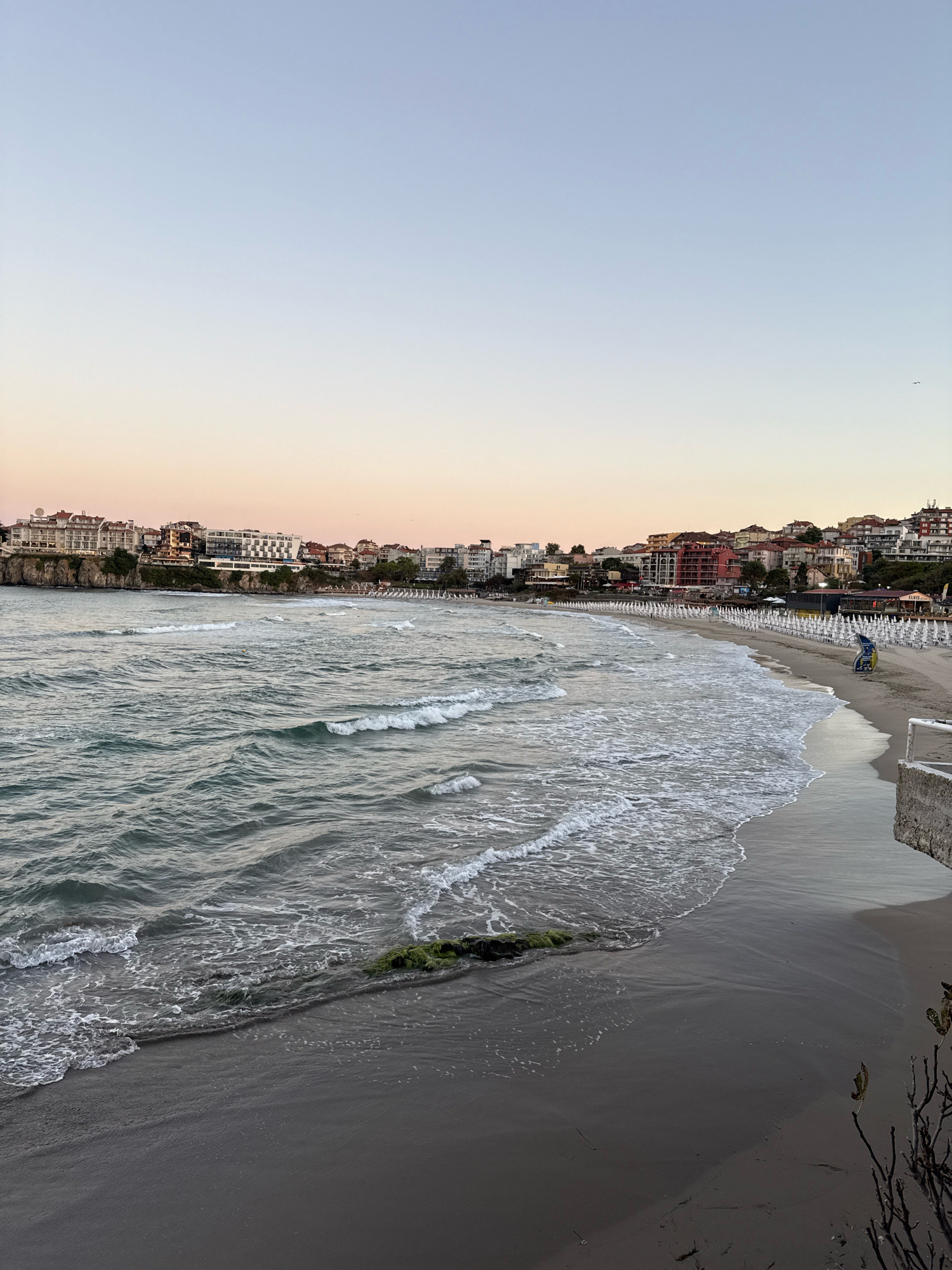 The image depicts a serene beach scene in Sozopol, Bulgaria, during the soft glow of sunset. The foreground is dominated by the gentle lapping of waves on a dark, sandy shore. To the right, a section of a concrete structure, likely part of a pier or promenade, juts into the frame, casting a shadow on the sand. Further along the coast, a long stretch of beach is lined with rows of white beach chairs and umbrellas, suggesting a popular tourist spot. Behind the beach, a hillside town rises, characterized by buildings with red-tiled roofs and a mix of architectural styles. The buildings are clustered closely together, creating a picturesque urban backdrop against the natural beauty of the sea and sky. The sky itself transitions from a pale blue at the top to a soft peachy hue near the horizon, indicative of twilight. A single person can be seen in the distance on the beach, adding a sense of scale and quiet solitude to the scene.