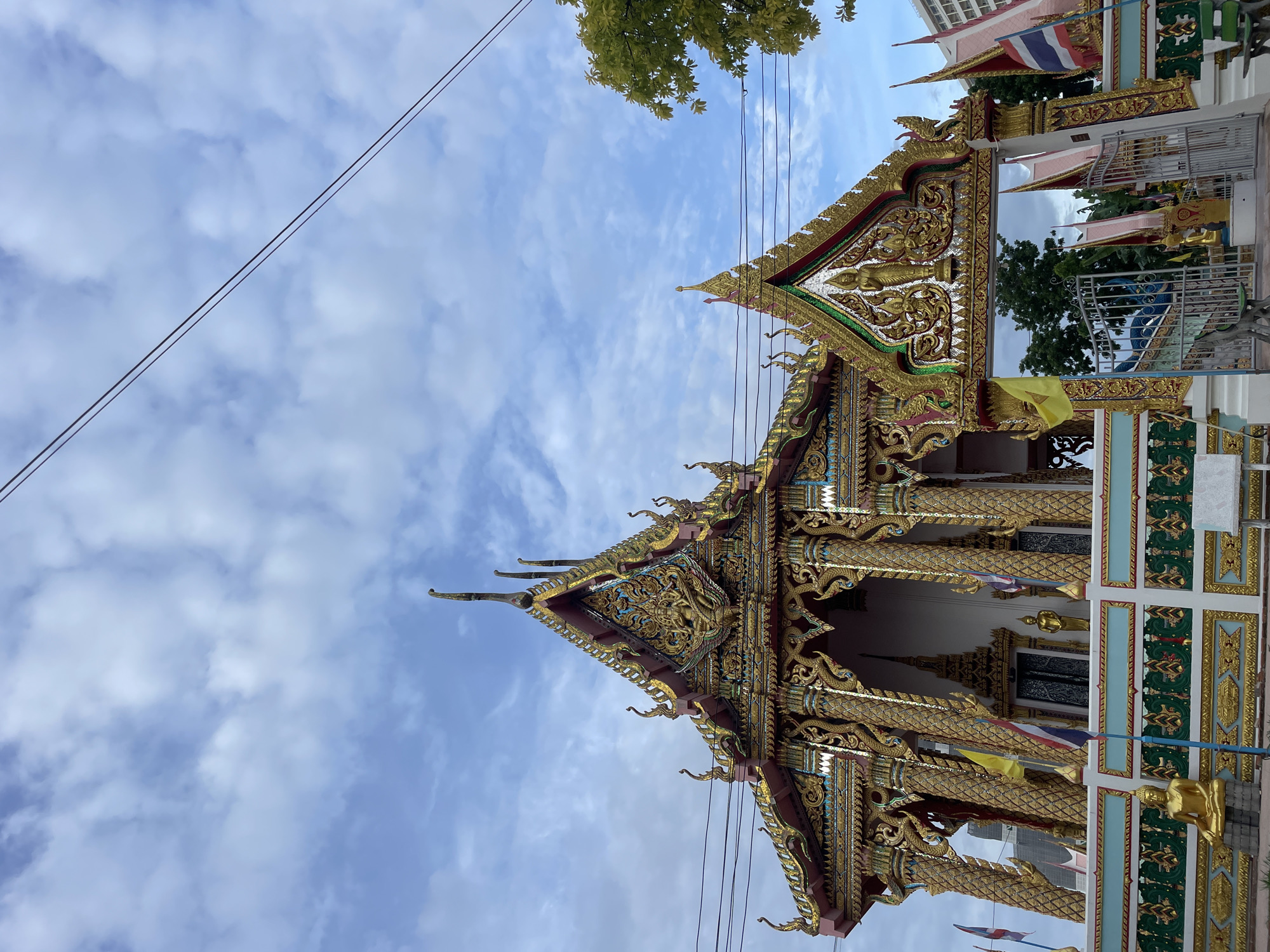 This is a photograph of the Wat Chai Mongkol Royal Temple, a Buddhist temple located in the city of Pattaya, Thailand. The temple is a prominent landmark in Pattaya, known for its ornate architecture and religious significance.
The image captures the main entrance to the temple, featuring a grand gateway adorned with intricate gold carvings and Buddhist iconography. The gateway is flanked by towering pillars and topped with a multi-tiered roof, characteristic of Thai temple architecture. Several flags, including the Thai national flag, are seen flying outside the temple.
The temple complex is surrounded by a decorative wall, also embellished with gold motifs. A statue of Buddha is visible in front of the wall, and other statues and religious artifacts are scattered throughout the grounds. The temple is set against a backdrop of a bright blue sky with scattered clouds, indicating a clear and sunny day.
The overall atmosphere of the photograph is one of serenity and spiritual reverence, reflecting the sacred nature of the temple. The image showcases the rich cultural heritage of Thailand and the beauty of its religious structures.