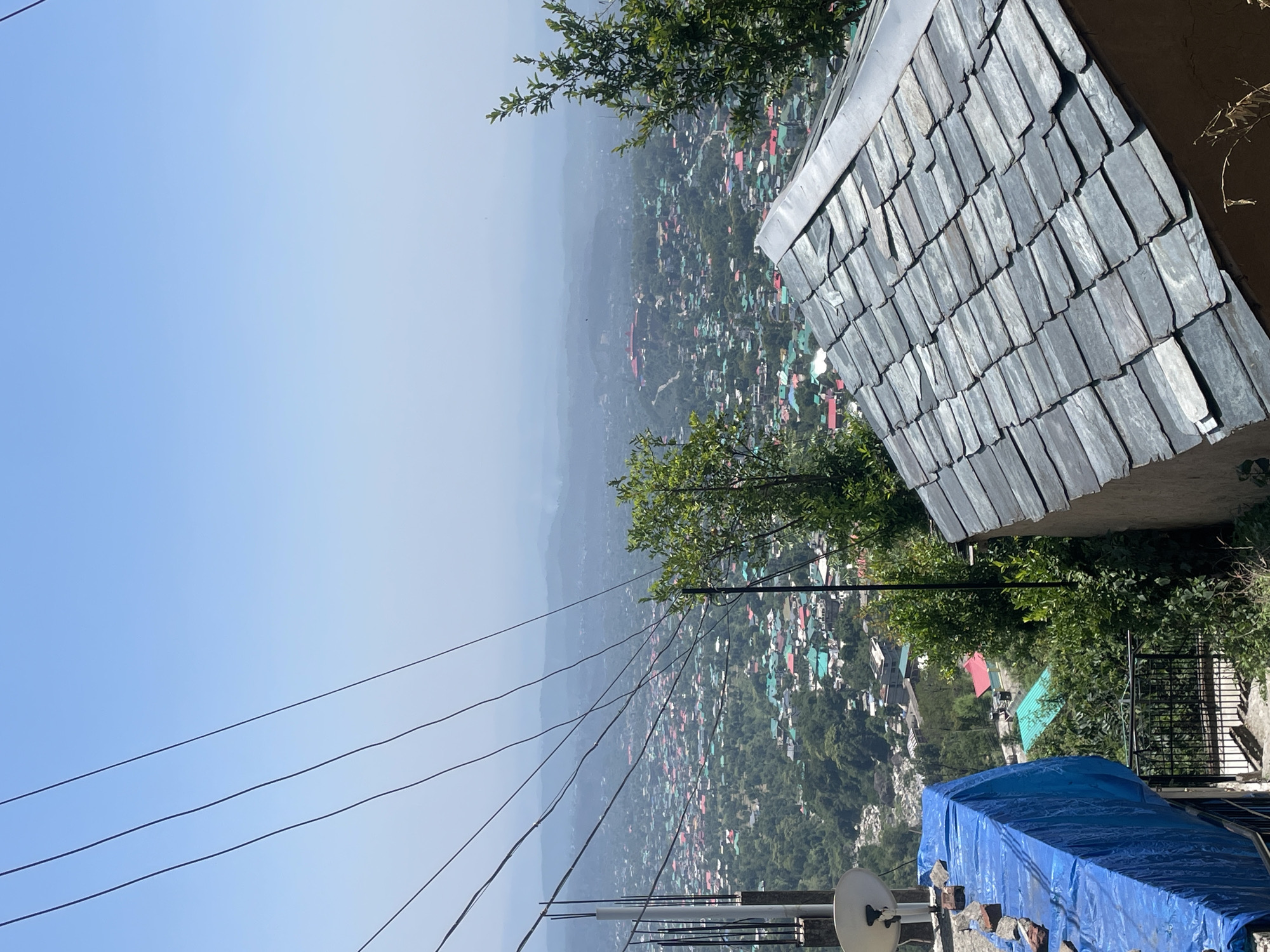 The image depicts a view overlooking a densely populated area in Dharamshala, India. The foreground features a slate-tiled roof, vegetation, and a blue tarp covering an object. Overhead power lines run across the frame, partially obscuring the view. The town below consists of numerous buildings with roofs of various colors (red, green, etc.), interspersed with trees. A misty haze obscures the distant mountains. The sky is a pale blue, transitioning to a lighter hue near the horizon. The presence of a satellite dish suggests modern communications infrastructure in the area.