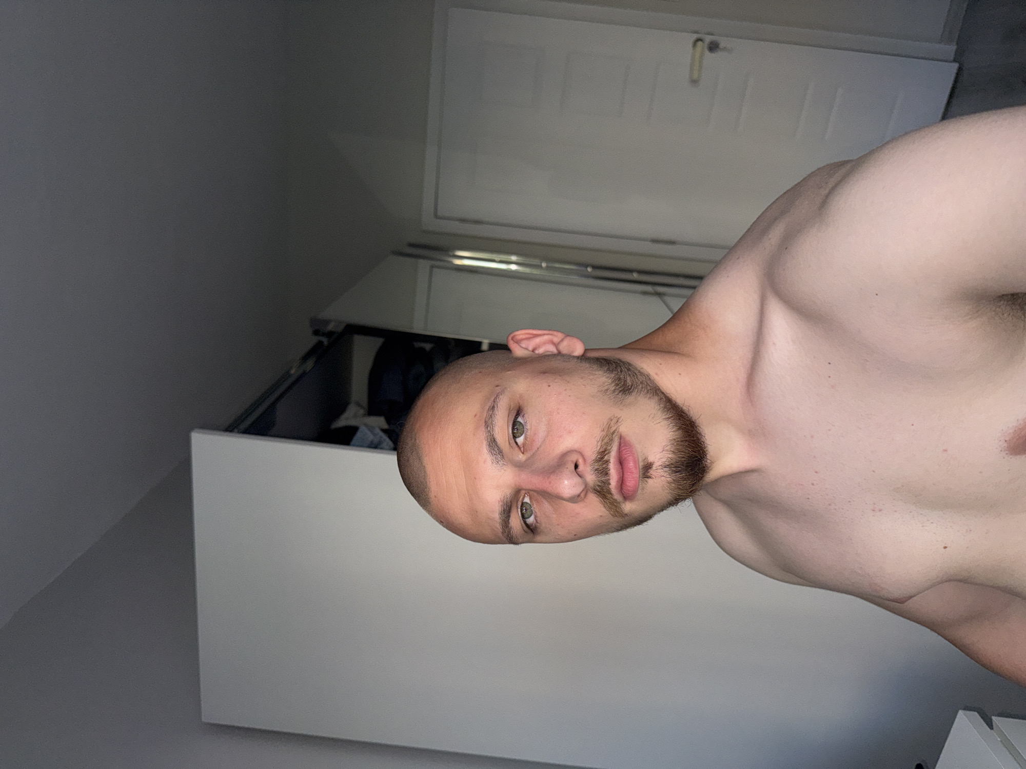 A young adult male, appearing in his 20s or early 30s, stands shirtless, facing the viewer directly. He has a shaved head, a goatee beard, and green eyes with a neutral expression. His upper torso, including his chest and shoulders, is visible, indicating a muscular build. The scene is indoors, in what appears to be a residential room in the city of Stara Zagora, Bulgaria. To the subject's left is a light gray or white wardrobe. To his right, a white door with a light-colored handle and keyhole is partially visible, with a narrow reflective surface, possibly a mirror or glass panel, positioned between the wardrobe and the door. The room is illuminated by soft, indirect light, casting subtle shadows.
