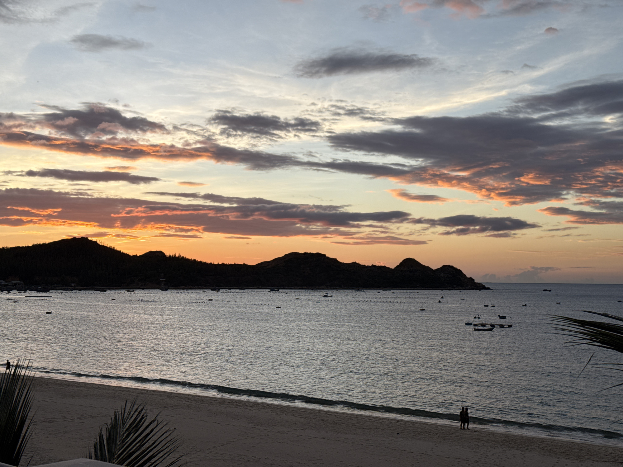 A serene beach scene unfolds during sunset, with a calm sea meeting a sandy shore. The sky is a canvas of warm orange and pink hues, painted by the setting sun, which is partially obscured by silhouetted mountains in the distance. Numerous small boats are scattered across the water, suggesting a fishing village or a popular seaside spot. Two figures, a couple, stand close together on the beach, appearing to watch the sunset. Palm fronds frame the lower right corner of the image, adding a tropical touch to the tranquil atmosphere. The scene evokes a sense of peace and natural beauty.