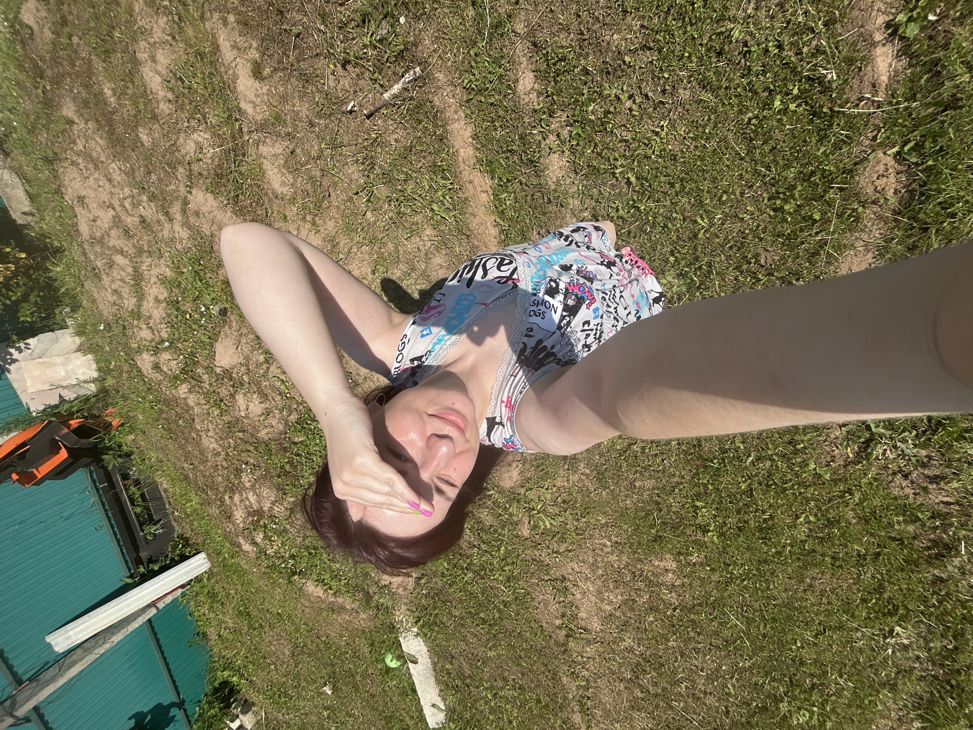 An overhead, high-angle selfie is taken outdoors by a person lying on the grass in the city of Foky, Russia. The individual, with brown hair and fair skin, is positioned on their back, with their right arm raised to shield their eyes from the sun. They are wearing a light-colored, sleeveless top with a colorful graphic print featuring text and abstract designs. The person's legs are extended, and their feet are partially visible.
The ground beneath is a mix of short green grass and dry, sandy soil. A white concrete slab is visible on the left, partially covered by grass. In the background, to the left, is a green corrugated metal fence and some overgrown vegetation. To the right of the fence, there are some industrial-looking structures with orange and black elements. The overall environment suggests a somewhat undeveloped or rural area, possibly a yard or a patch of land adjacent to a residential or industrial zone. The sunlight is strong, casting shadows on the ground.