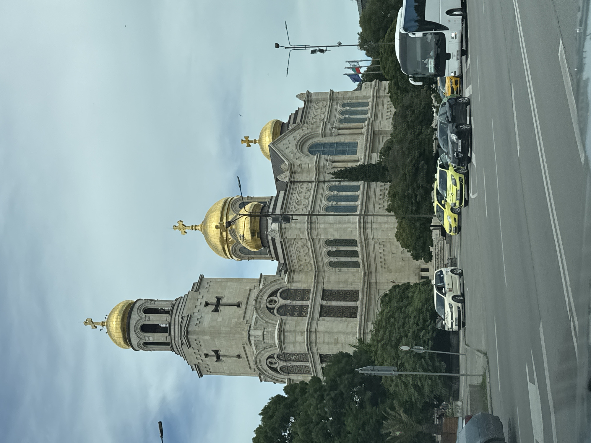 This image, taken in Varna, Bulgaria, captures a view of the Cathedral of the Assumption of the Virgin, a prominent landmark with distinctive golden domes and ornate architecture. The cathedral is partially visible, with its imposing facade and religious iconography. In the foreground, a street with white lane markings runs parallel to the building. Several vehicles are present on the road, including a white bus at the top, a black car, and a couple of yellow taxis. The scene is set against a cloudy, overcast sky. Trees are visible on the left side of the frame, providing a backdrop of greenery. The overall impression is one of an urban setting with a significant religious structure, likely during daylight hours given the ambient light. The orientation of the image is vertical, as if captured from a moving vehicle. No specific activity or event is clearly depicted, suggesting a general street-level view. The image is undated and there are no explicit emotional cues conveyed. There is some visible text on the white bus, which appears to be a destination or route indicator.