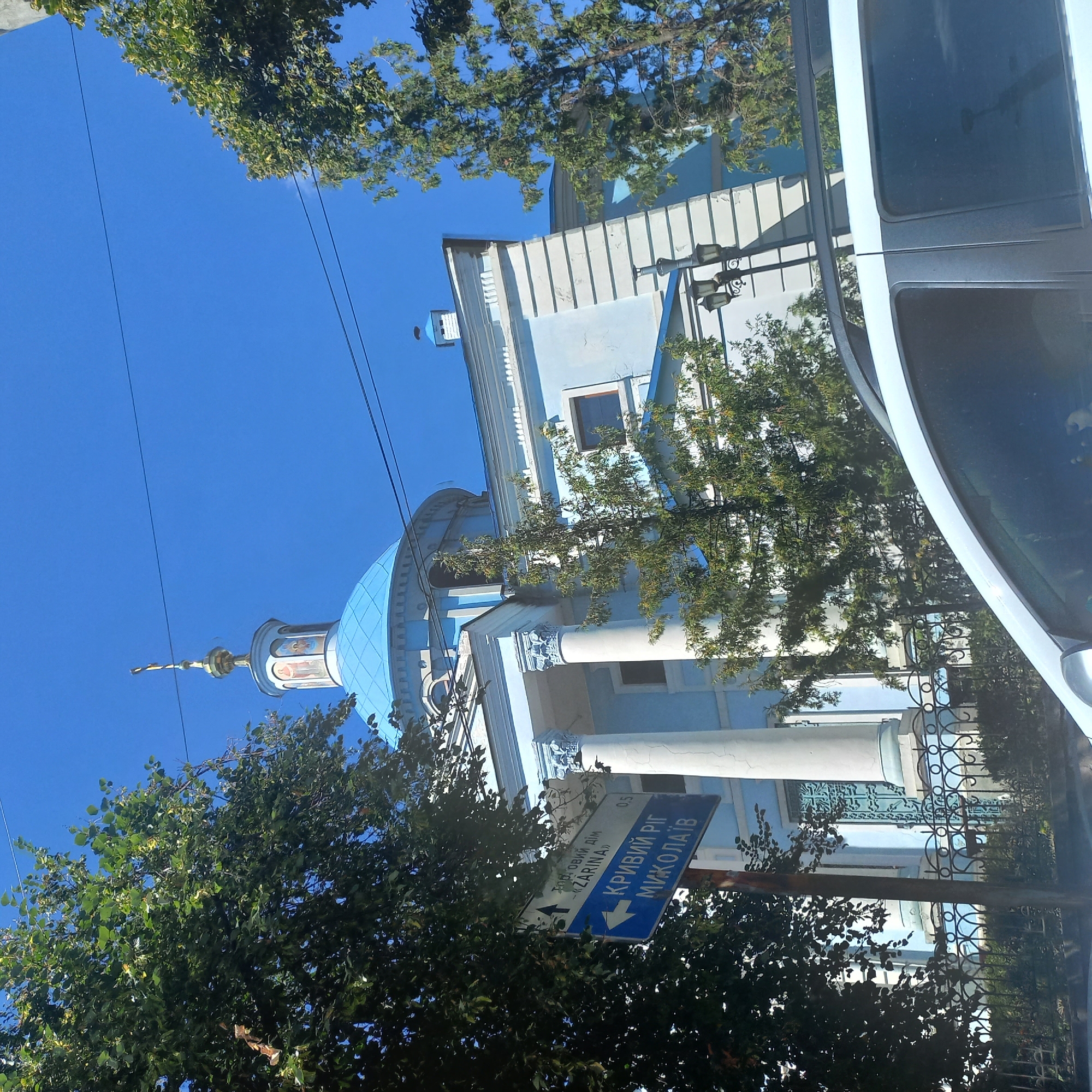 This image depicts a street scene in Kropyvnytskyi, Ukraine, featuring a light blue building with a prominent blue dome, likely a church or administrative building, against a clear blue sky. A signpost is visible on the left, with text in Ukrainian indicating directions to "TORGOVYI DIM - 'ZARINA'" and "KRIVYYI RIG MYKOLAYIV". Several trees with green foliage partially obscure the building. A portion of a white vehicle is in the foreground, suggesting the photo was taken from within or next to a car. The architectural style of the building, with its columns and decorative elements, is characteristic of Eastern European religious or public structures. The overall impression is one of a sunny day in an urban environment.