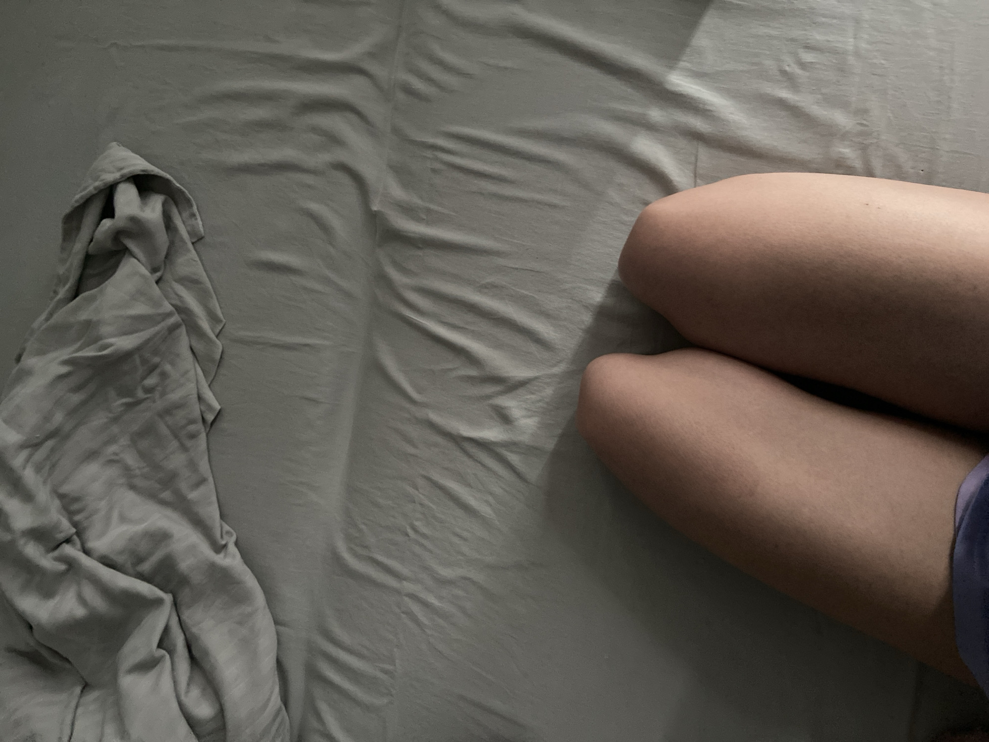 The image depicts a person's legs and lower torso lying on a bed with gray sheets. The sheets are rumpled and appear to be made of a soft, possibly linen or cotton material. The lighting suggests it is either early morning or late evening, with a warm glow casting shadows across the bed. The person is wearing purple shorts. The scene is intimate and domestic, with no other people or objects visible. There is no information in the image to suggest a specific location in Minsk, Belarus.