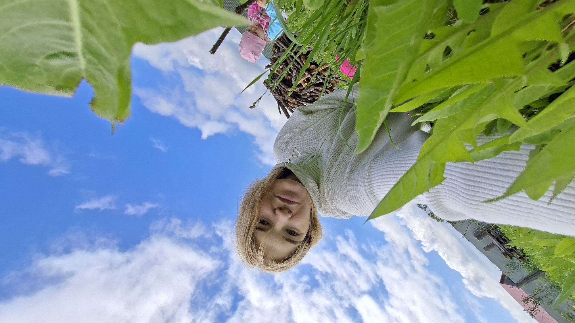 The image presents an outdoor scene captured from a very low angle, looking upwards towards the sky. In the foreground, a blonde woman with shoulder-length hair and light-colored eyes looks directly at the camera with a neutral expression. She is wearing a light-colored, possibly off-white or light gray, ribbed turtleneck or collared sweater, featuring a small triangular metal detail on the zipper pull.
Behind the woman, and partially visible through green foliage, is a small child wearing a pink hat and possibly blue clothing. Further back, a pile of neatly stacked small wooden branches or logs is seen. The entire scene is framed by vibrant green leaves and grass, suggesting a garden or natural outdoor environment.
Above the subjects, a clear blue sky filled with scattered white, fluffy clouds dominates the background. On the far right side of the image, a building with a reddish roof and what appears to be a dark-colored wall is visible in the distance. The bright lighting indicates it is daytime, with fair weather. The photograph captures a casual moment outdoors, possibly a selfie from a unique perspective within the vegetation. No visible text is present in the image. The location context provided is Большой Букор, Russia.