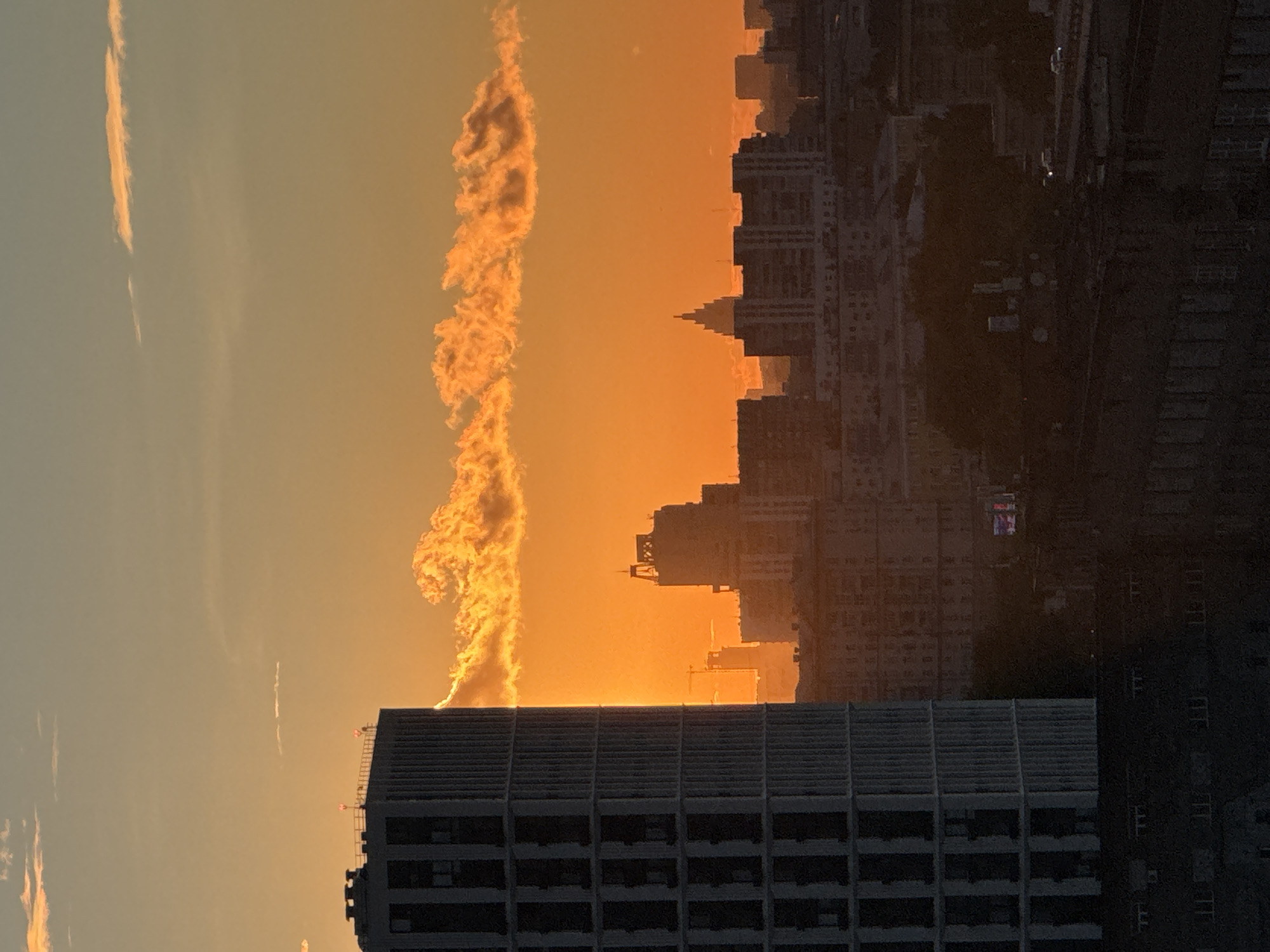 The image shows a cityscape at sunset in Moscow, Russia. A tall, cylindrical building with a patterned facade dominates the left side of the frame, partially obscuring a vibrant orange sky illuminated by the setting sun. A line of clouds, lit from below by the sun, stretches across the mid-ground. In the background, silhouetted buildings of varying heights form the urban landscape, with a distinctive spire visible on one of the central structures. The lower portion of the image depicts darker, more densely packed buildings, suggesting a lower elevation or older part of the city. The scene conveys a sense of urban sprawl bathed in the warm, dramatic light of dusk.