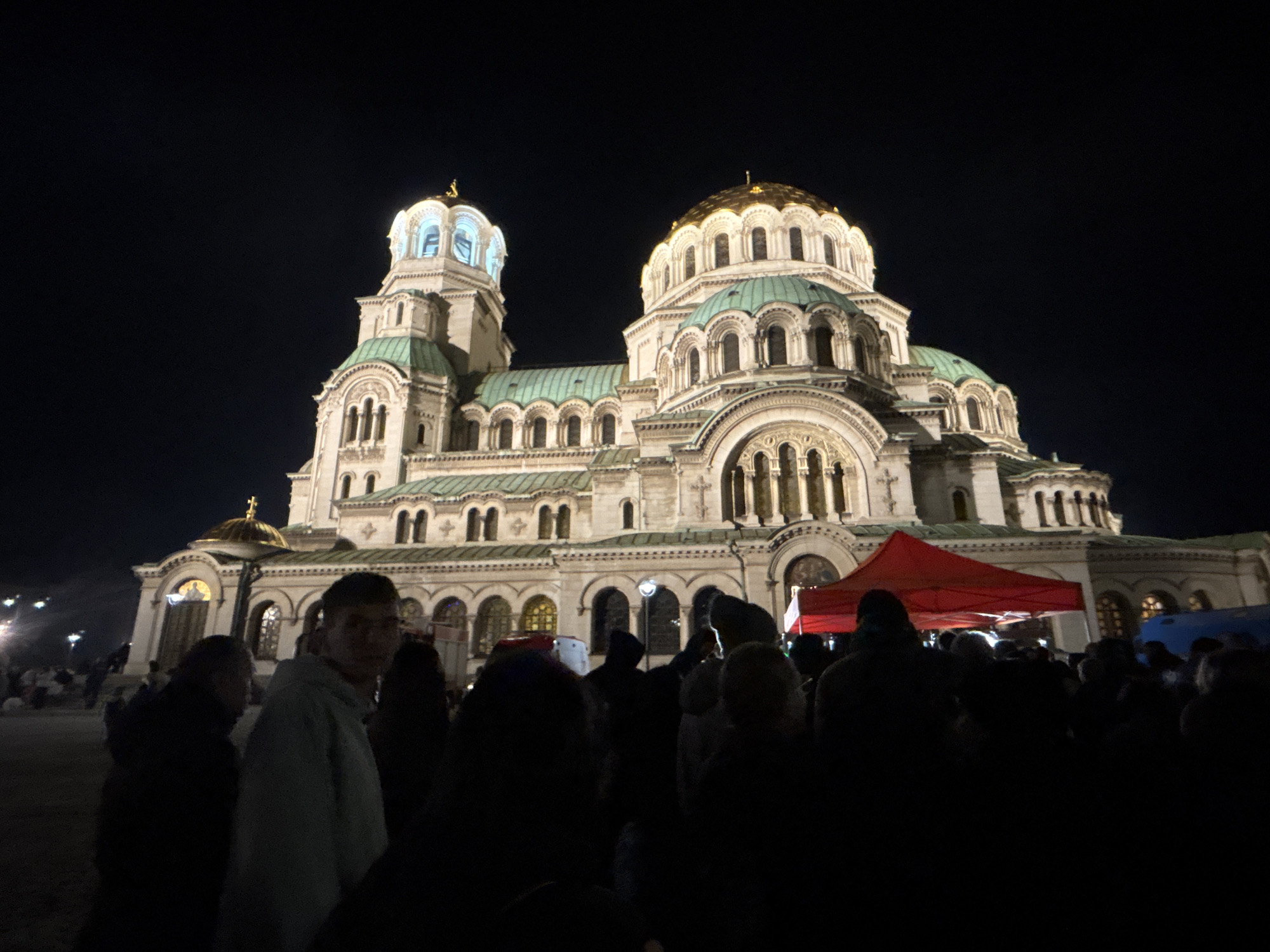 At night, a large, ornate church is illuminated, standing behind a crowd of people. A red tent is visible among the crowd. One person in the crowd is facing the camera. The church has multiple domes, one with a golden top. The location is most likely Alexander Nevsky Cathedral in Sofia, Bulgaria.