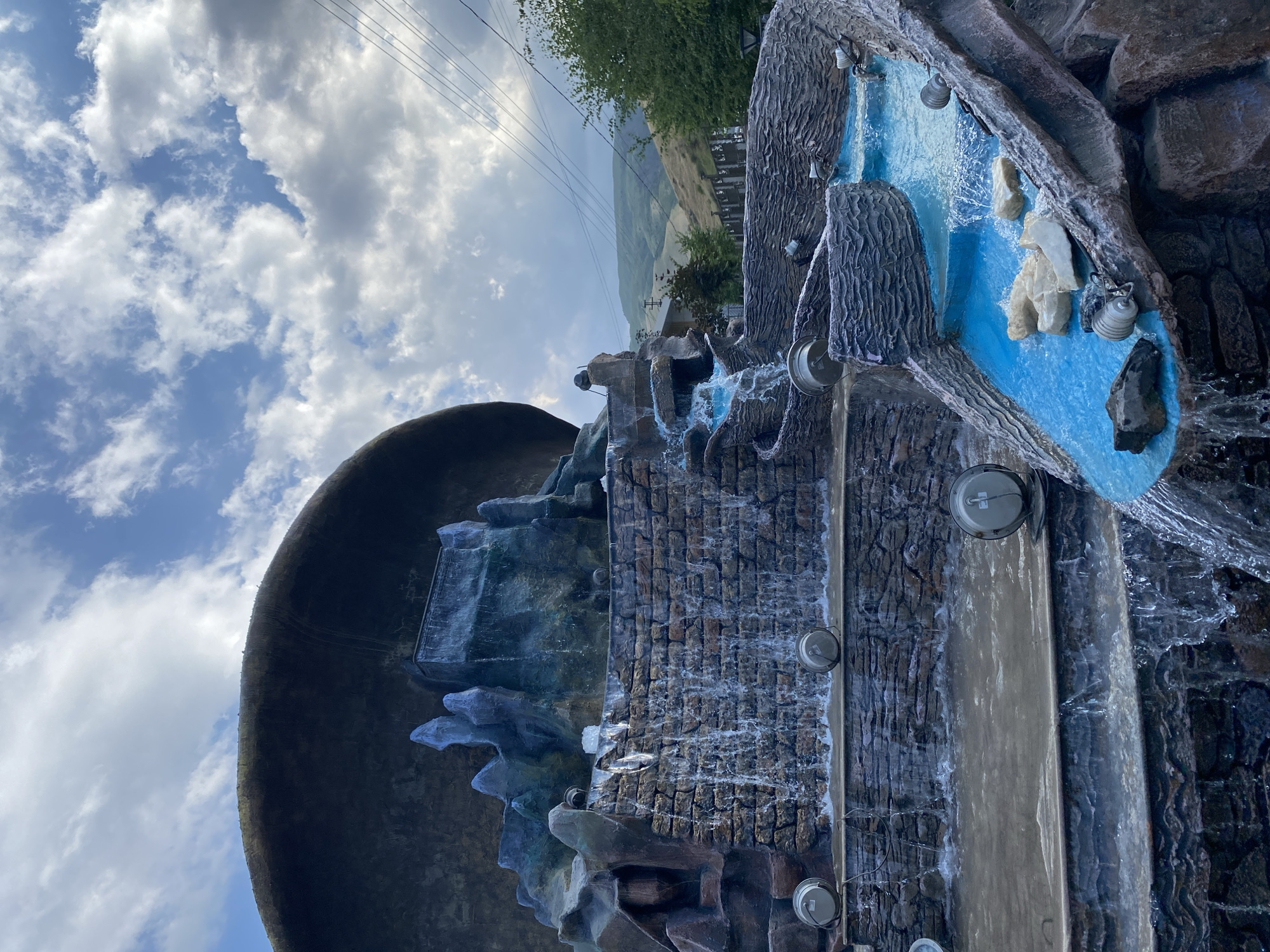 This image shows a multi-tiered artificial waterfall structure located in Arashan, Kyrgyzstan. The waterfall is constructed with stone-like materials and features blue-tinted water flowing down various levels. Several round, metallic lights are visible along the structure, some submerged in the water. The background reveals rolling hills and a cloudy sky. There are no people visible in the image. The overall scene depicts a man-made water feature set within a natural landscape.