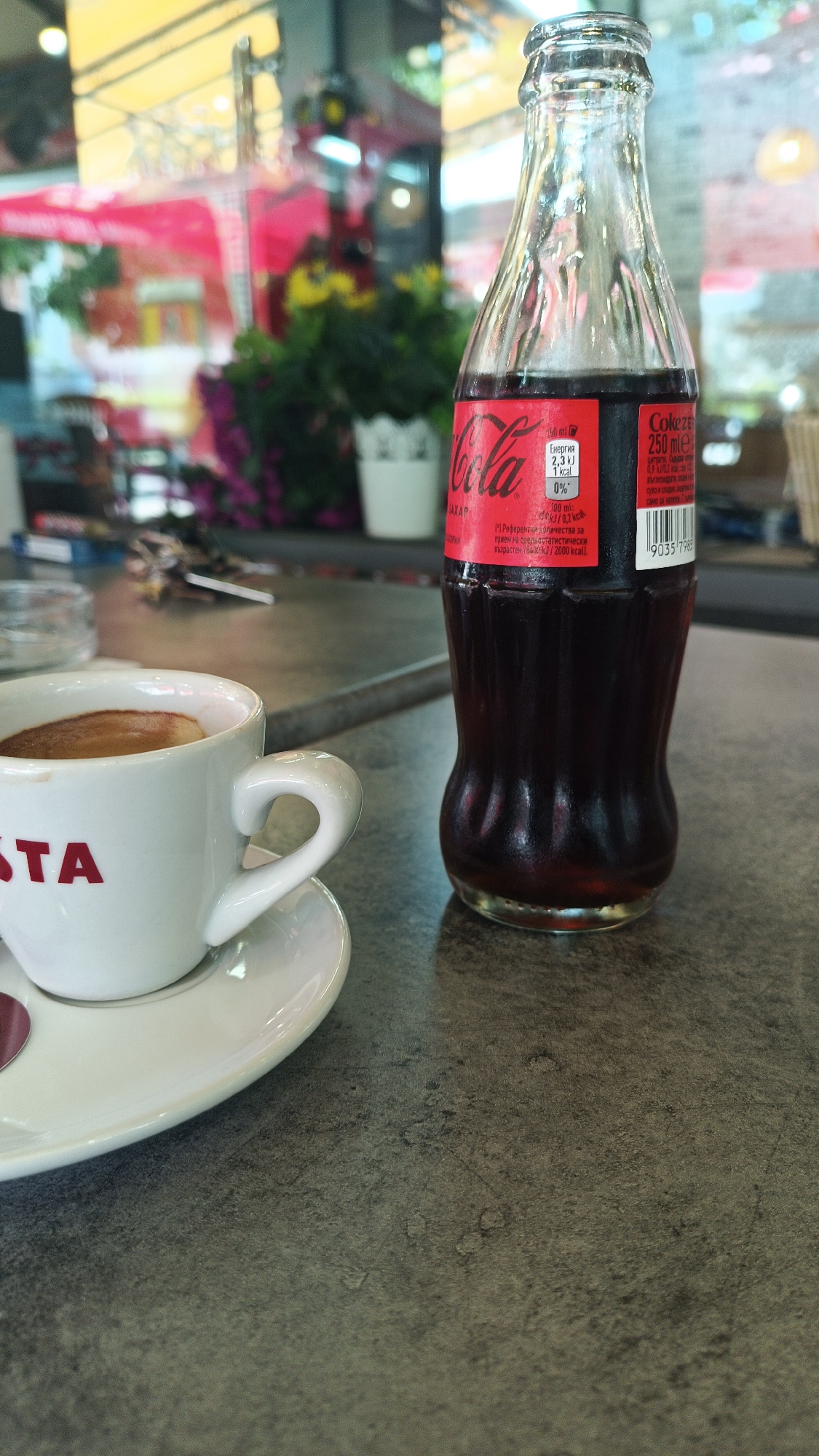 On a textured gray surface, a white Costa coffee cup sits on a white saucer next to a small glass Coca-Cola bottle. The cup is positioned on the left, while the bottle stands upright to the right. Behind the objects, a blurred background reveals an outdoor setting with plants and indistinct structures, possibly a café or restaurant setting. The OCR text on the bottle suggests the image was taken in Bulgaria, and based on the Costa coffee, potentially in the city of Karnobat. There are no people visible in the frame.