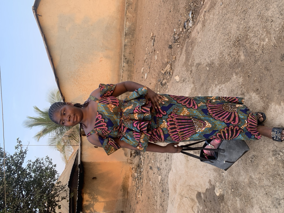A woman stands outdoors, wearing a colorful, patterned dress with off-the-shoulder details. She holds a large, dark-colored tote bag. The background features a dirt ground and buildings with varied roofing materials, under a bright, clear sky. A palm tree is partially visible to the left of the woman, and other trees are present.