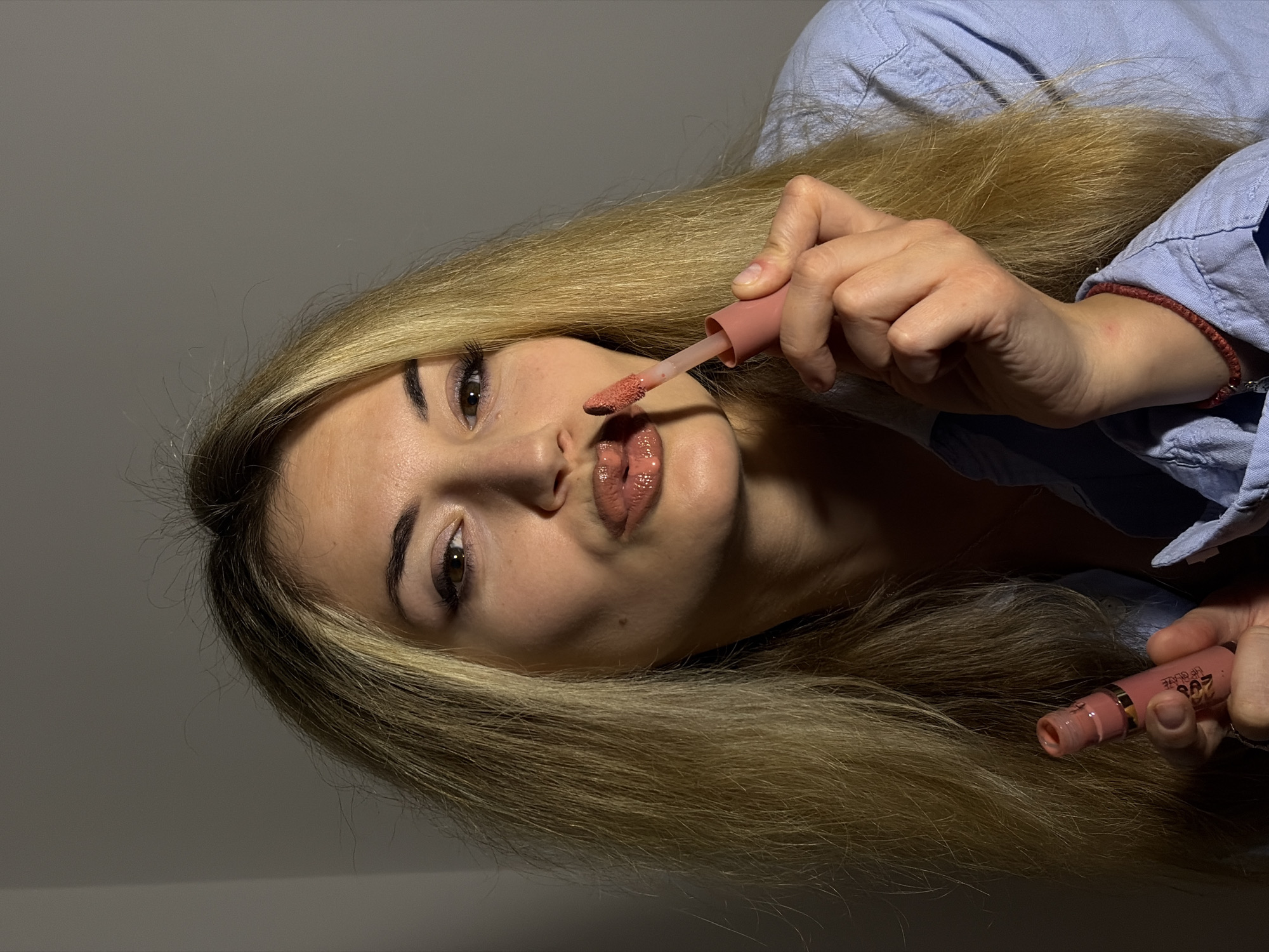 An overhead shot, rotated 90 degrees clockwise, captures a young woman applying lip gloss in an indoor setting in Budva, Montenegro. She has long, straight blonde hair that falls around her face and shoulders. Her features include dark eyebrows, long eyelashes, and brown eyes, which are directed towards the camera. Her lips are slightly parted and puckered as she uses a small applicator wand from a light pink lip gloss tube. The lip product she is applying appears to be a glossy, peachy-pink shade.
In her right hand, she holds the applicator wand, while her left hand holds the main body of a second, similar lip gloss tube. On the tube in her right hand, the text "LIP GLOSS" and a number resembling "208" are clearly visible. She is dressed in a light blue, collared shirt, and a thin, red string or woven bracelet is noticeable on her right wrist. The background consists of a plain, light gray wall or surface. The lighting is soft and directional, highlighting her face and hair. The overall scene depicts a focused moment of personal grooming.