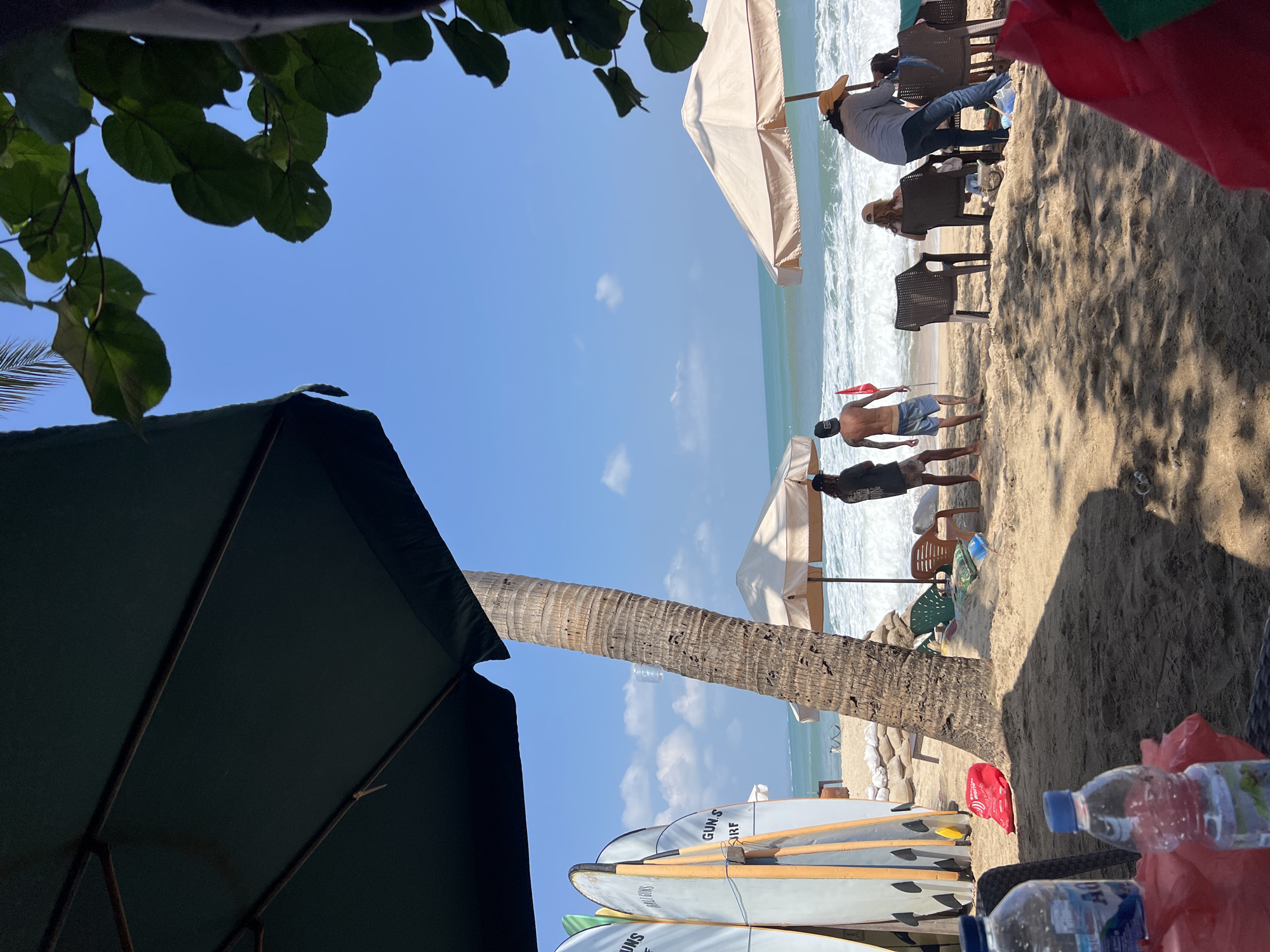 The image depicts a beach scene in Kuta, Indonesia, on a sunny day. In the foreground, two plastic water bottles sit on a table covered with a red cloth, casting shadows on the sand. To the left, a row of surfboards stands upright, leaning against a palm tree trunk. Beyond the surfboards, beach umbrellas and lounge chairs are scattered on the sand. Three people are visible near the water's edge, two men standing and one person seated. The ocean waves are crashing onto the shore. In the background, the sky is clear and blue, with a few white clouds. The overall impression is a relaxed beach atmosphere.