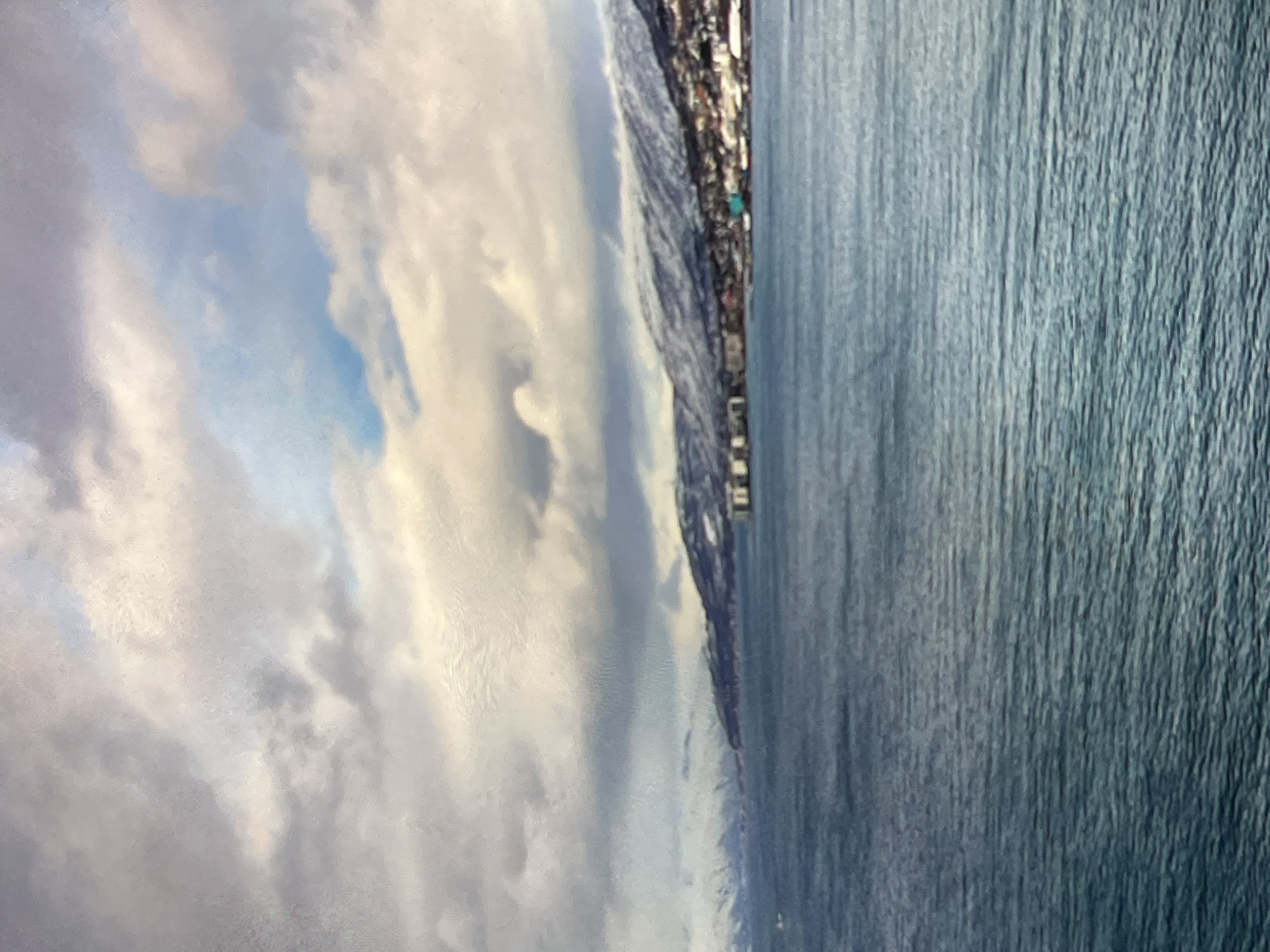 The image shows a body of water with a mountainous, snow-capped coastline in the background. A town or city is visible along the coast, nestled at the base of the mountains. The sky is filled with dramatic, textured clouds. The water is a deep blue-green, with small ripples visible on its surface. The overall impression is of a vast, natural landscape with human settlement in proximity.